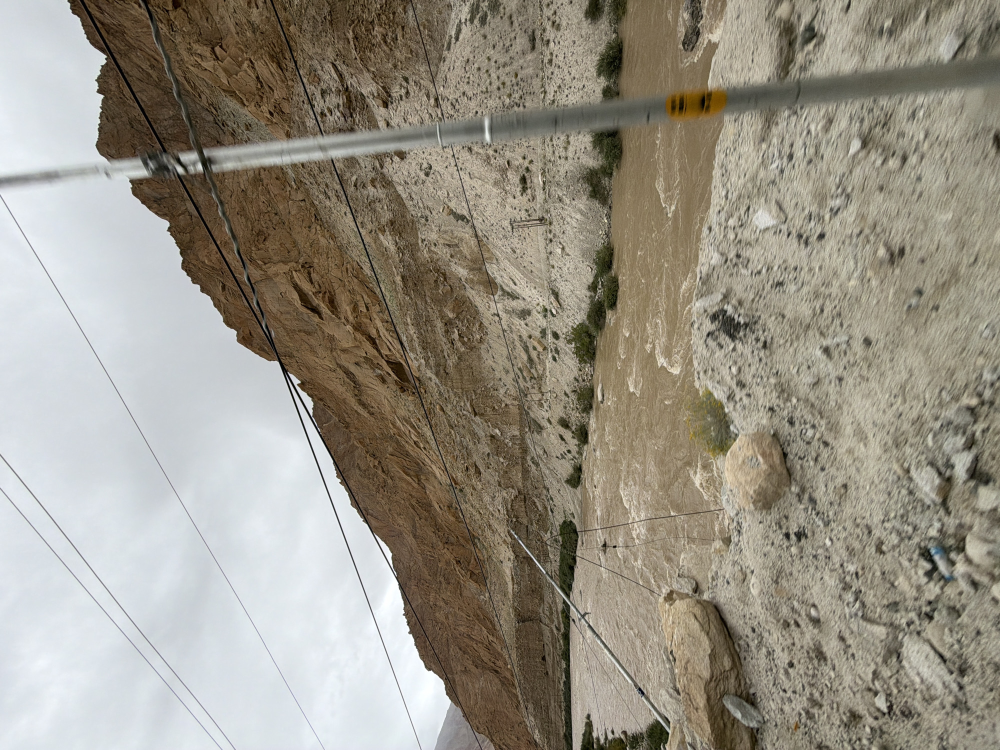 The image depicts a wide, muddy river flowing through a rocky, mountainous gorge. The river is turbulent with visible currents and eddies. The banks of the river are characterized by loose scree and sparse vegetation. In the foreground, the ground is composed of sand, gravel, and scattered rocks. A metal pole with a yellow marker is present on the right side of the image, obscuring part of the view. Several power lines stretch across the scene, supported by poles, indicating infrastructure in the area. The sky is overcast and gray. The scene is likely located in a remote, arid region, possibly Ladakh, India, given the geological formations and arid landscape often found there. There are no people visible in the image.
