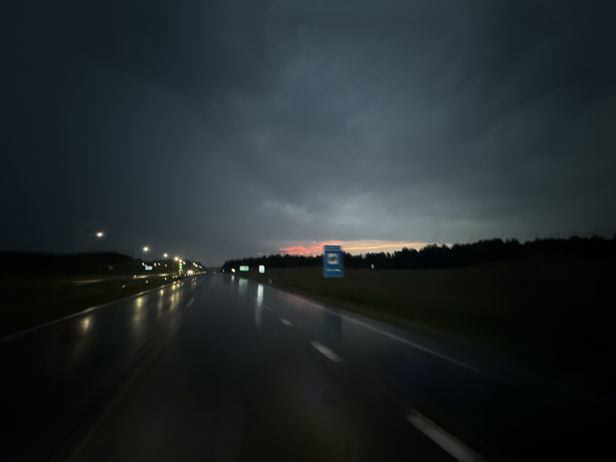 The image depicts a dark, moody scene of a wet road at dusk or night, likely on a highway. The road surface is highly reflective, showing streaks of light from streetlights and possibly headlights. In the distance, there is a hint of a colorful sunset or twilight sky peeking through dark, cloudy conditions. The roadside features a dark treeline on the right and what appears to be the edge of a field or undeveloped land on the left, with some distant lights. A blue sign with an image is visible on the right side of the road. The overall impression is one of a drive through a somewhat desolate, atmospheric landscape under challenging weather conditions. The specific location in Драчкаўскі сельскі Савет, Belarus, is not visually identifiable from the image itself.
