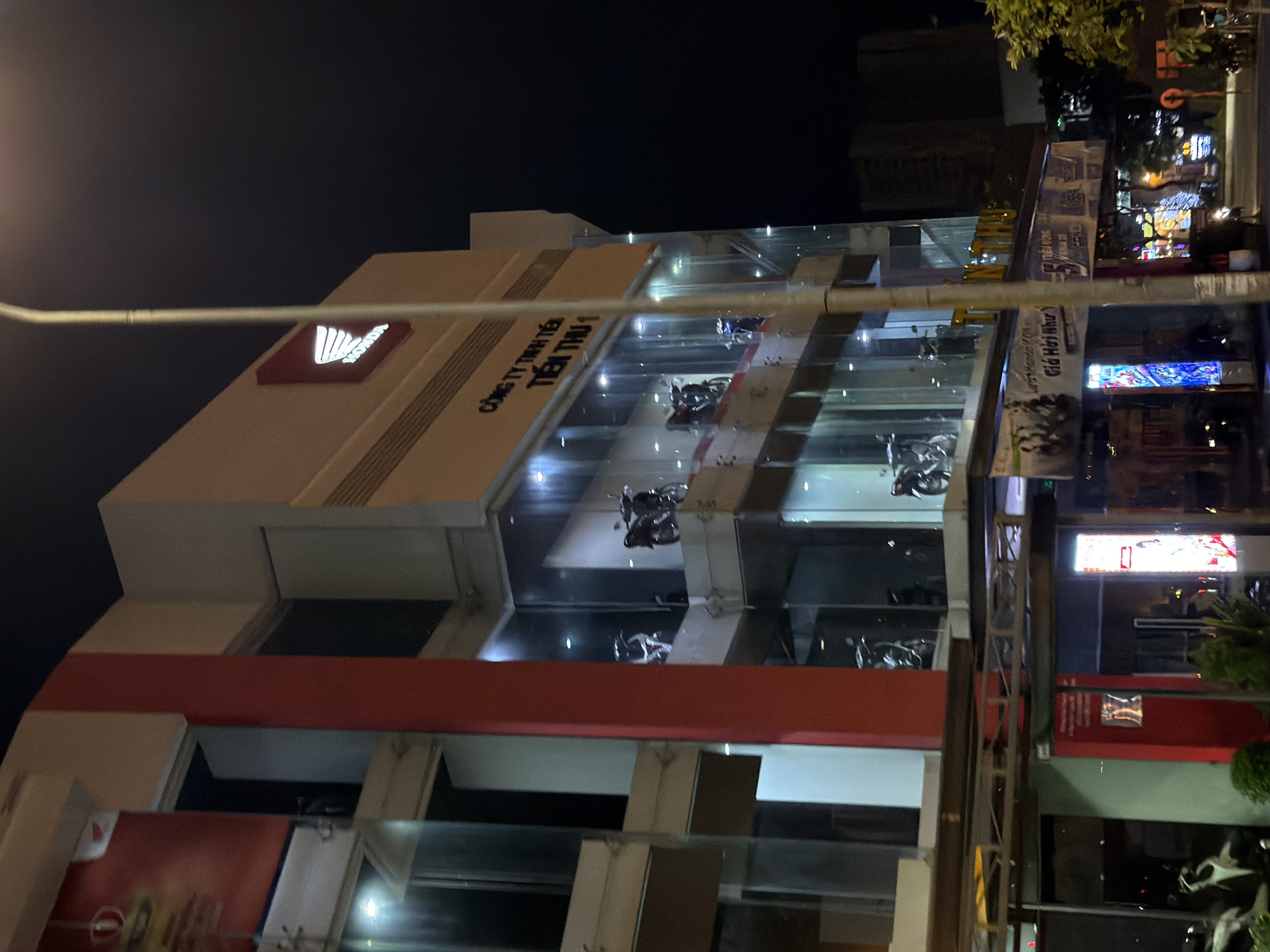 This is a nighttime photograph of a multi-story building in Đà Nẵng, Vietnam, likely a Honda dealership. The building has a modern design with large glass windows showcasing motorcycles on display on multiple levels. The exterior is lit, and the Honda logo is visible, a red diamond shape with white wings and the word "HONDA" in white lettering. Text in Vietnamese is present on the building, including "CÔNG TY TNHH TIẾN THU 1" which translates to "Tien Thu 1 Co., Ltd.". Banners with Vietnamese text and images of people and motorcycles are also visible at street level. The environment appears to be an urban setting, with streetlights and other illuminated signs in the background. No people are clearly visible or identifiable in the image.