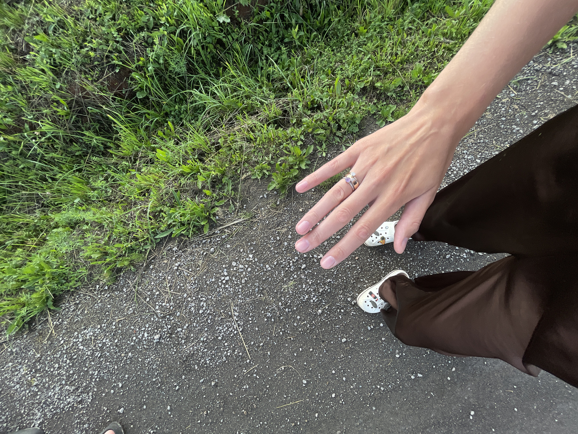 The image shows a person's legs and hand from a first-person perspective. The person is standing on a gravel path adjacent to a grassy area. The person is wearing dark brown trousers and white Crocs with cartoon characters. The person's right hand is extended towards the camera, revealing two rings on the ring finger. The background consists of lush green grass, suggesting an outdoor, possibly rural or park-like setting. The overall lighting indicates it is daytime, likely late afternoon or early evening due to the soft light. There is no specific indication of the location being within the city of Podshivalovo, Russia, as the image lacks any identifiable landmarks or context clues specific to that location.