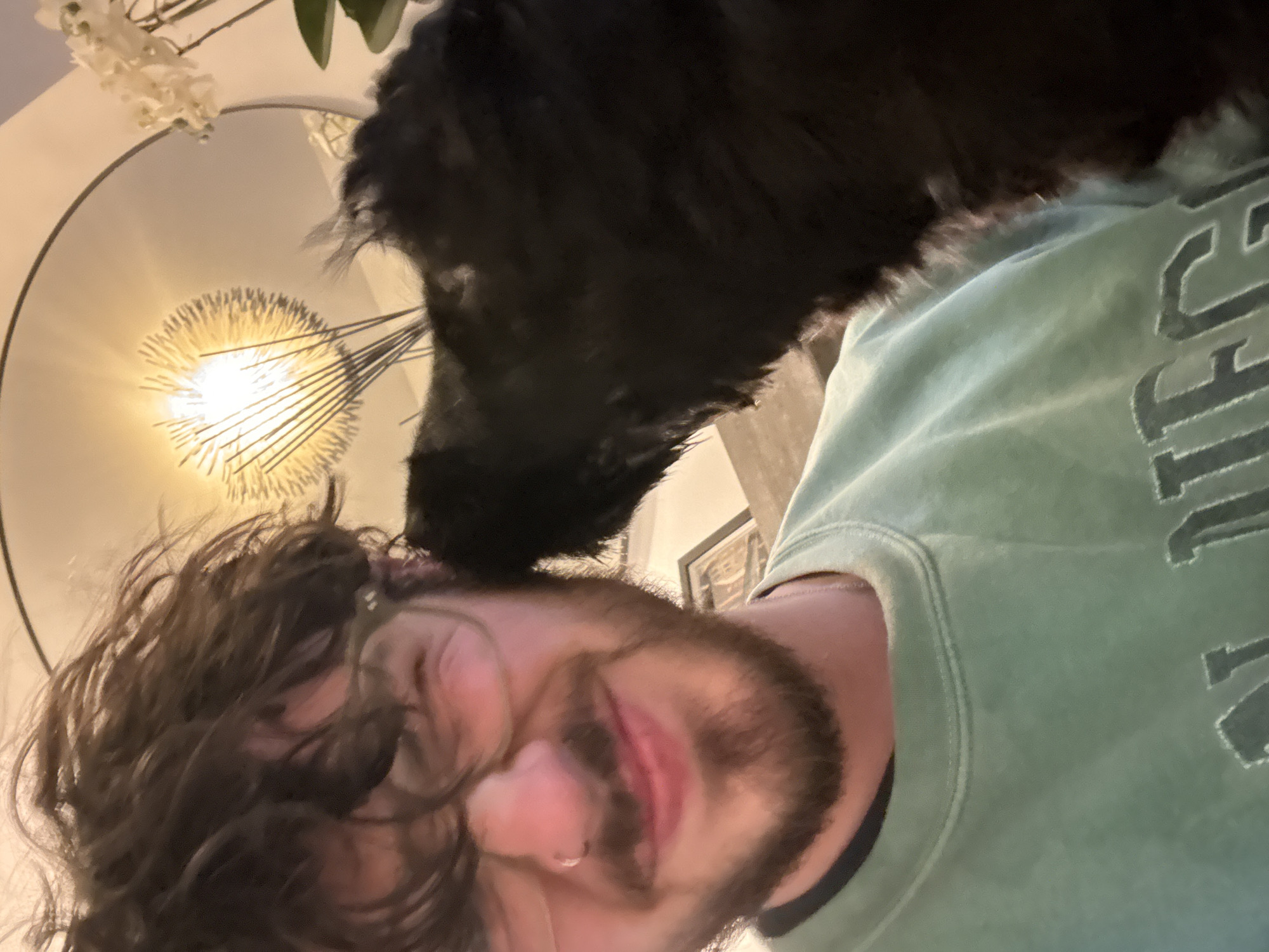 A close-up shot shows a man with curly brown hair and a beard. He is wearing a green sweatshirt. A black dog is nuzzling the top of his head. A circular light fixture with radiating elements is visible in the background. The presence of the light fixture and floral decoration, suggests this scene is taking place indoors. The city and location are unknown.