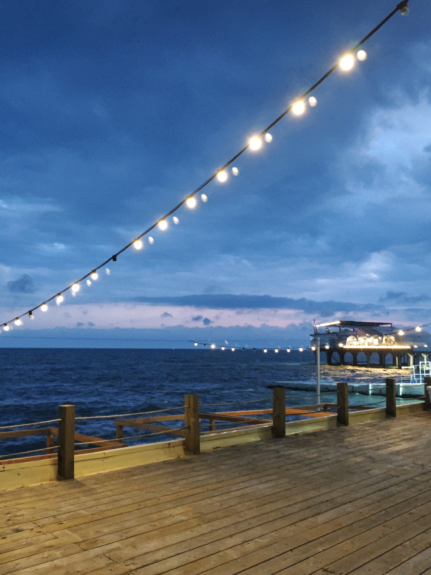The image depicts a coastal scene in Sochi, Russia, at dusk or twilight. A wooden pier extends into the sea. The pier is illuminated by overhead string lights. A dark sea separates the pier from a building structure. The sky is a mix of blue and grey. The pier railing is wooden with rope accents. The planks of the pier are also wooden, with visible grain. The lights above the pier glow brightly against the darkening sky. The water is slightly choppy. The building structure, possibly a restaurant or entertainment venue, is also illuminated with lights along its facade, and is partially built over the water.
