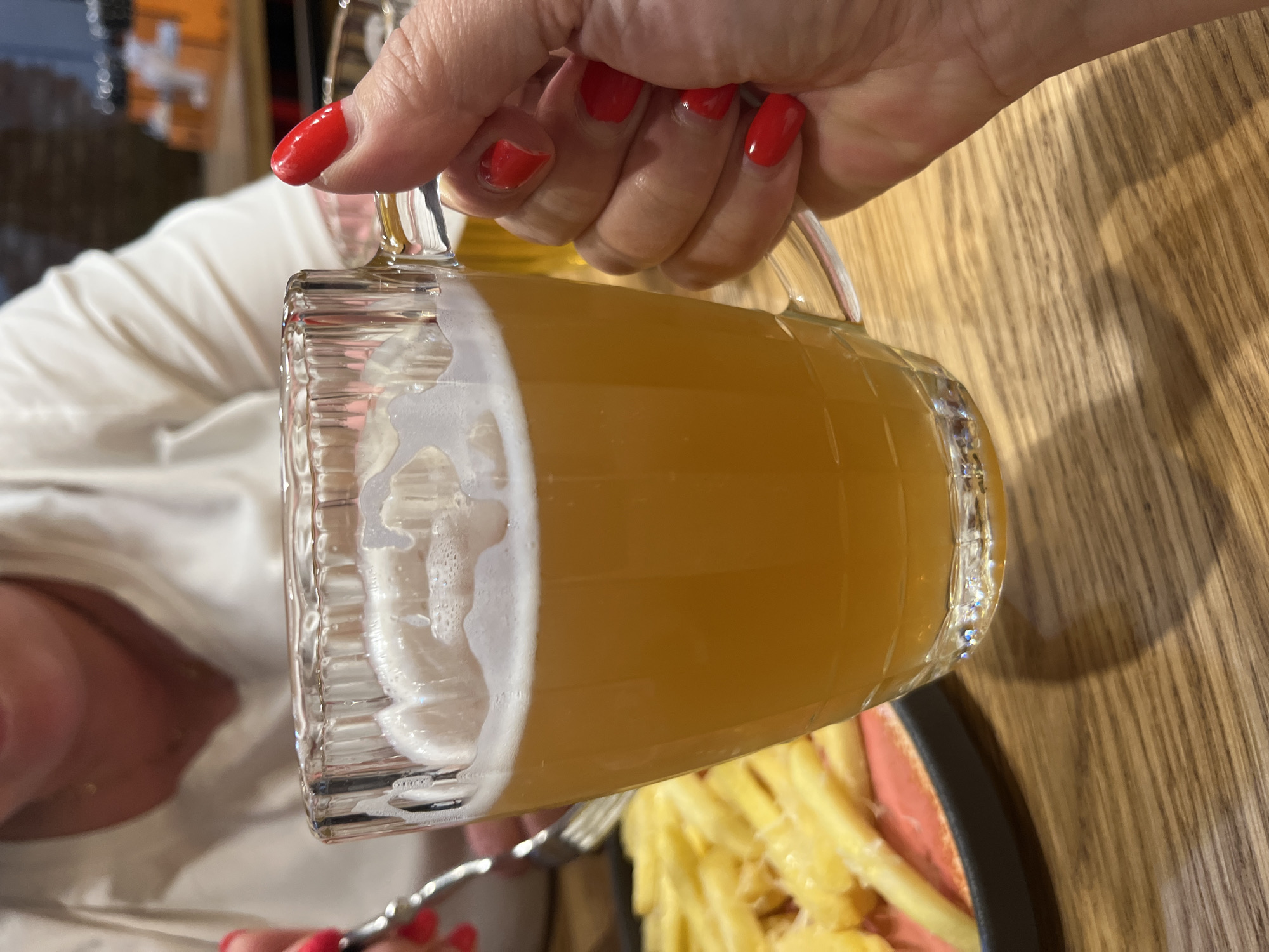 A close-up shot focuses on a hand with bright red nail polish gripping a clear glass mug filled with a pale orange beverage and a frothy white head. In the background, a portion of a person wearing a white shirt is visible, along with a plate of golden french fries. The scene appears to be set at a wooden table, possibly in a restaurant or bar. The location is identified as Novo-Nikolskoye, Russia.