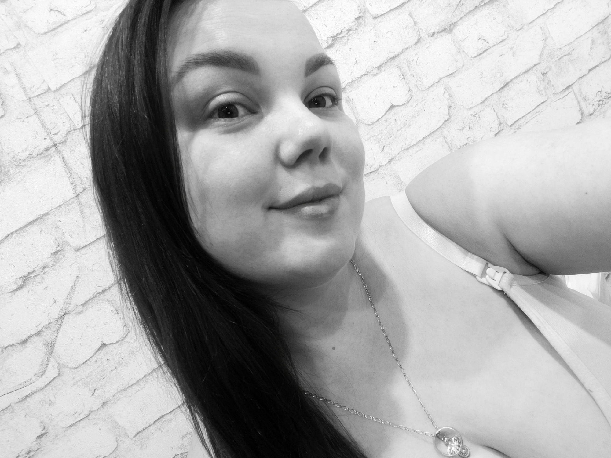 A close-up, black and white photograph shows a woman with dark hair and fair skin. She is wearing a necklace with a pendant. Part of a white garment with straps is visible on her shoulder. The background is a brick wall. The image does not contain sufficient information to confirm its location in Chaikovsky, Russia.