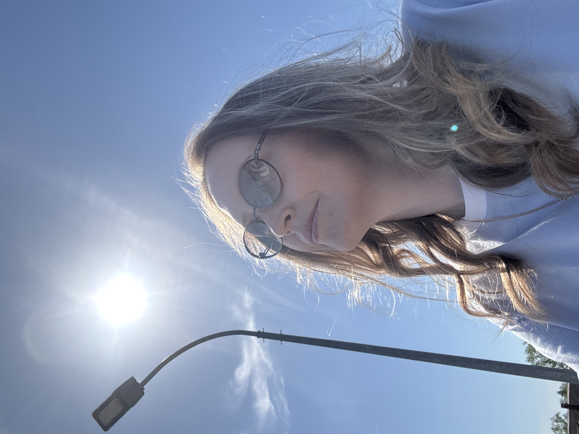 A woman wearing sunglasses and a light blue top is photographed from a low angle against a bright blue sky with the sun visible. A street lamp is also in the frame. The location cannot be determined as Balezino, Russia from the provided image.