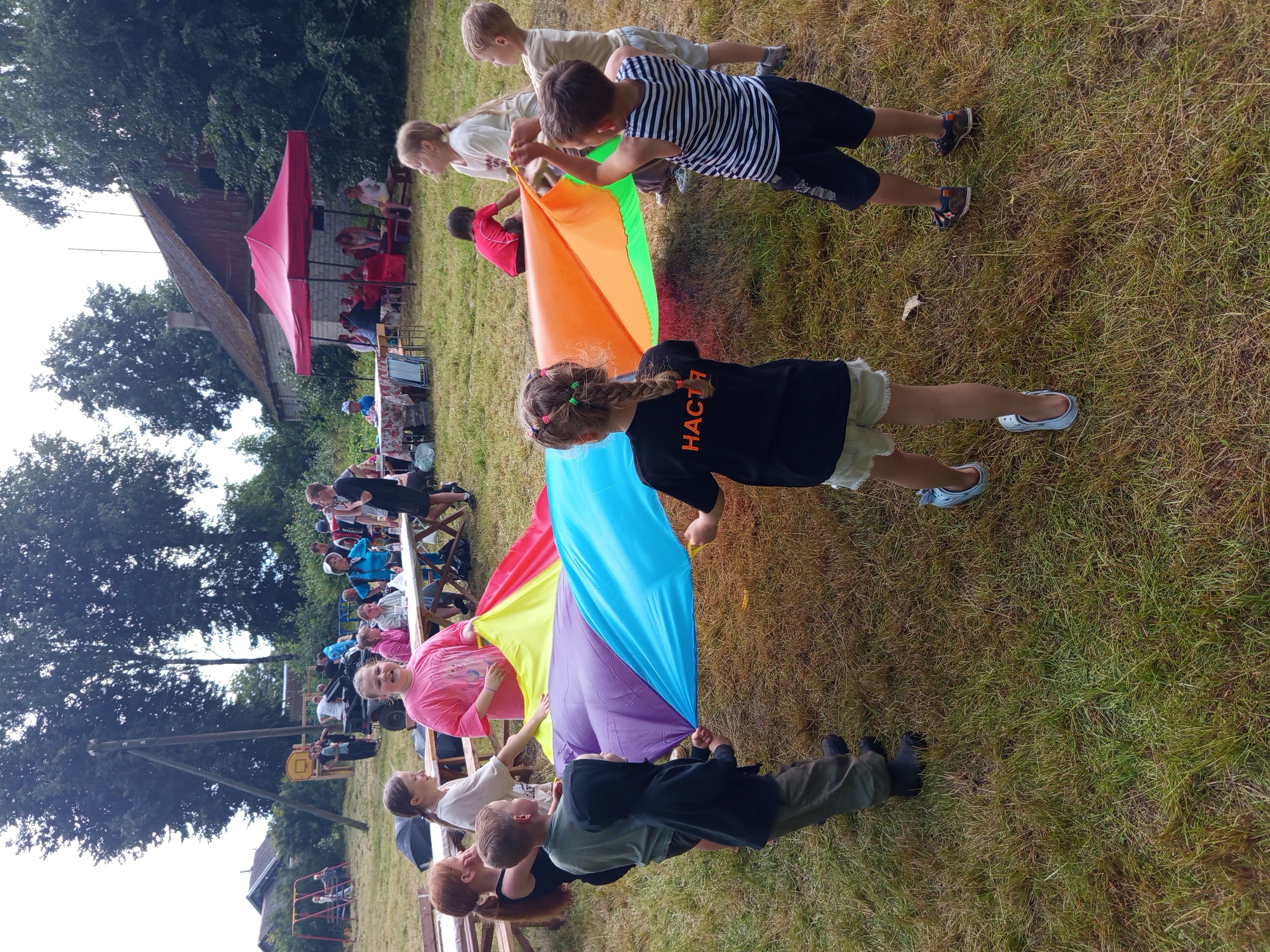 A group of approximately ten children and adults are gathered in an outdoor grassy area, participating in a game with a large, multi-colored parachute. The parachute is spread out on the ground, with children holding onto its edges, creating a dome-like structure. In the background, there are trees, a wooden structure that resembles a playground, and a building with a red roof. A red canopy tent is set up, and several tables with people seated around them are visible. The scene appears to be part of an outdoor event or festival, possibly in the city of Ostrovtsy, Russia, given the presence of what looks like a typical Russian rural landscape. The sky is overcast.