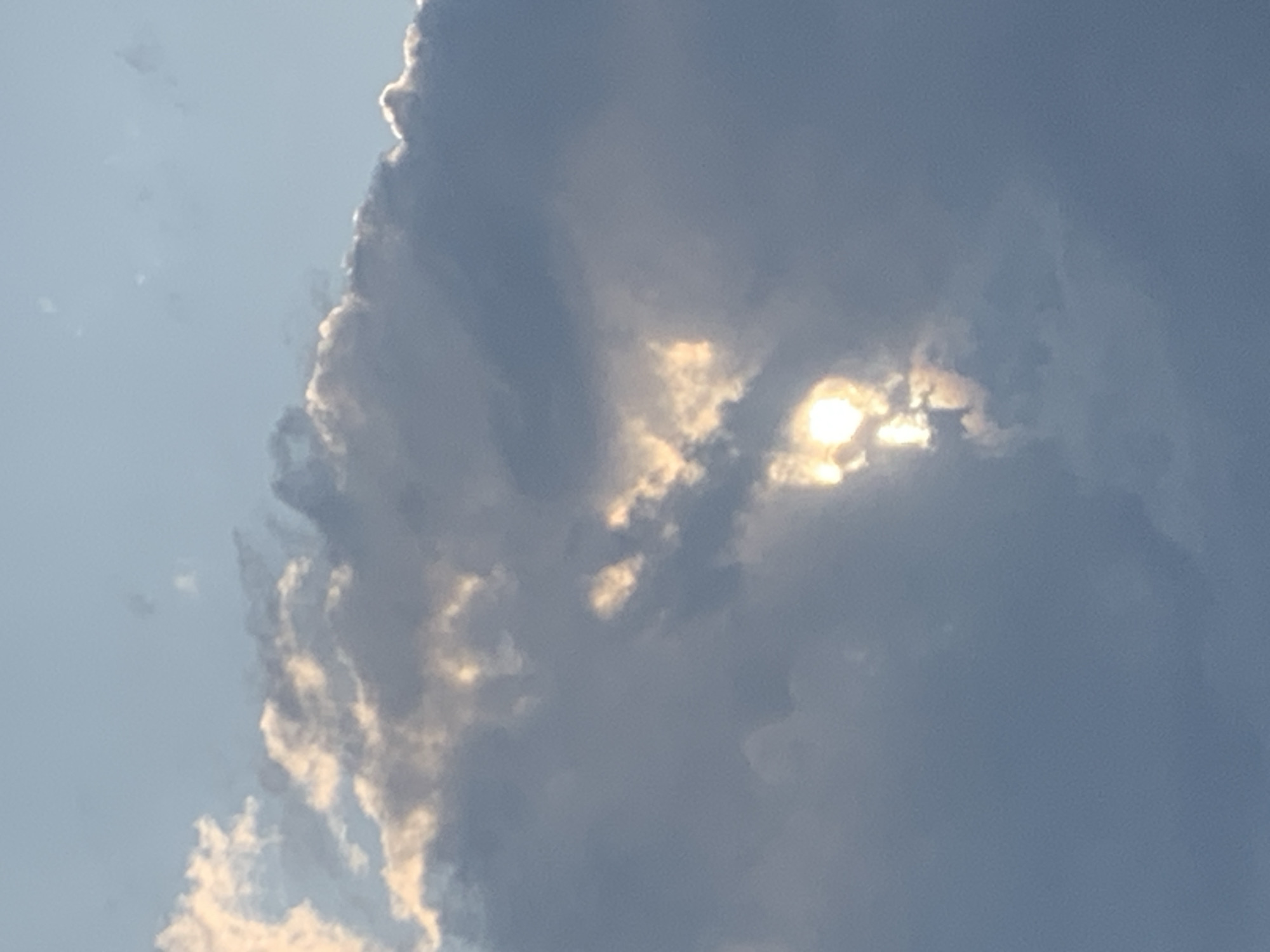 The image depicts a close-up view of clouds in the sky, with the sun shining through a gap. The clouds are a mix of dark gray and lighter white and yellow hues, suggesting they are illuminated from behind by the sun. The sky is a clear blue. The sun appears as a bright, concentrated light source peeking through the clouds, casting rays that create a luminous effect. There are no discernible people, buildings, or specific landmarks visible, and therefore, no identifiable location within the city of Kolomyia, Ukraine. The scene is a natural sky formation.