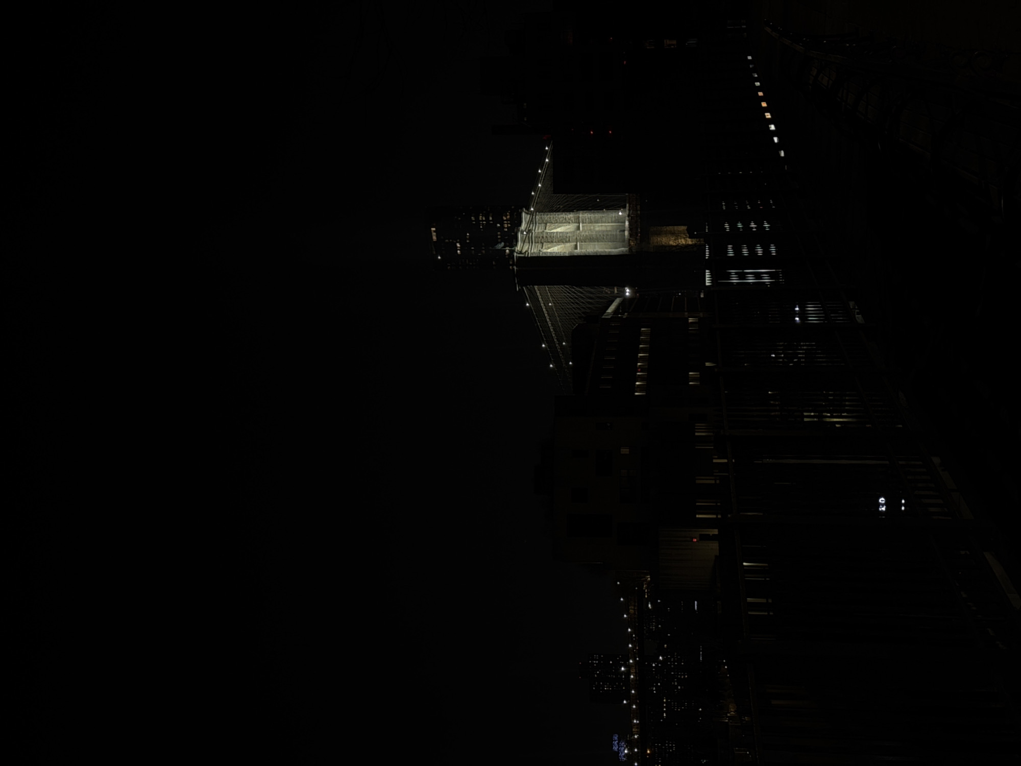 The image depicts an aerial view of the Manhattan Bridge at night in New York City. The bridge's structure, including its towers and cables, is illuminated. Adjacent to the bridge, the lighted windows of buildings line the streets of the city. The scene is predominantly dark, with the bridge and city lights providing the main sources of illumination.