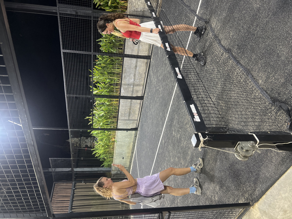 This image depicts an outdoor padel tennis court at night in Pecatu, Indonesia, illuminated by artificial lighting. Two adult females are present on the black-surfaced court, which features white boundary lines and is enclosed by black wire mesh walls. A black net, bearing "THE GROUND" logo, divides the court, with a winch mechanism visible on one of its support posts.
The female on the left has blonde hair tied back and is dressed in a light purple sports bra, matching shorts, light blue socks, and grey athletic shoes (New Balance visible). She carries a grey tote bag on her left shoulder and holds a smartphone in her right hand, pointed forward as if capturing an image or video. She is looking towards the right side of the frame.
The female on the right has dark, curly hair and is wearing a red spaghetti strap top, a white pleated skirt, and black athletic shoes. She holds a black padel racket in her right hand, resting it on the net, and a black smartwatch is visible on her left wrist. She is facing left, appearing to look towards the first female.
In the background, beyond the court enclosure, a concrete planter filled with lush green tropical foliage is visible. Behind the plants, a traditional Balinese stone shrine is partially discernible against the dark night sky.