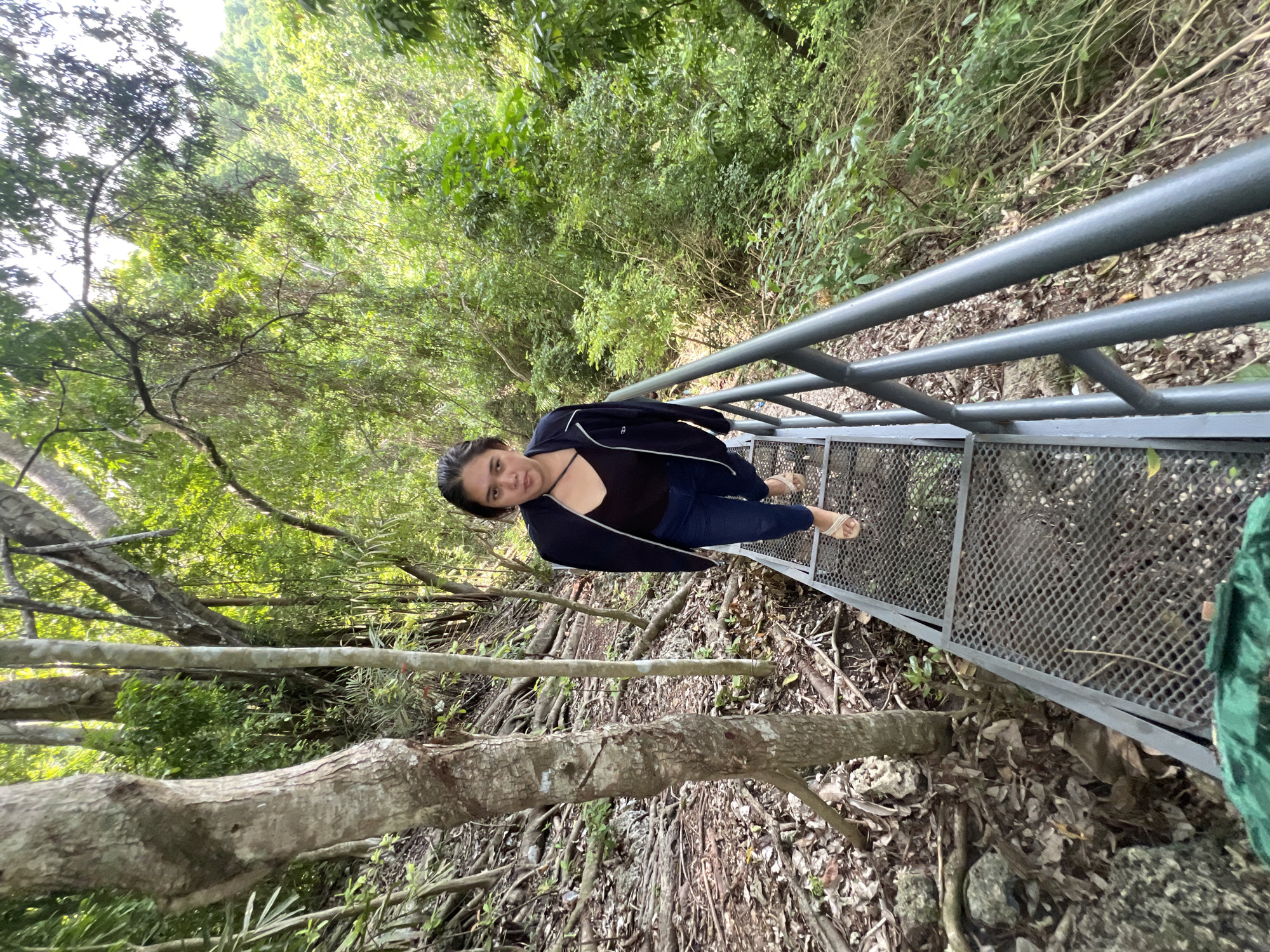 The image shows a woman walking on a metal walkway in a lush, green, tropical environment. The walkway, made of interconnected metal grates, is lined with a dark metal railing. The woman is wearing a dark jacket over a black top and blue jeans, and she has on sandals. She is looking towards the camera with a neutral expression. The walkway is positioned on a sloping path surrounded by dense vegetation, with trees and foliage filling the background. The ground beside the walkway is covered with fallen leaves and exposed roots, indicative of a natural, forested area. The lighting suggests it is daytime, with sunlight filtering through the canopy. Based on visual cues commonly associated with Samal Island, Philippines, such as the tropical flora, the presence of elevated walkways for navigating natural terrain, and the humid, verdant atmosphere, this scene is likely located in a nature park or a tourist attraction on Samal Island, Philippines.