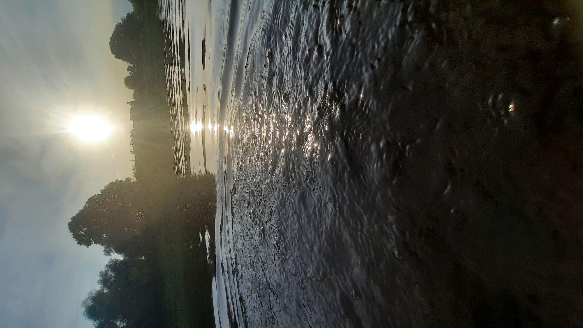 The image depicts a body of water, possibly a river, with a bright sun setting or rising in the background. The sun's rays are casting a glare on the water's surface, creating a shimmering effect. Trees and a faint silhouette of buildings are visible on the far bank, suggesting a rural or semi-rural setting. The foreground is dominated by the dark, rippling water, with some small bubbles or disturbances visible. The overall impression is one of natural tranquility.