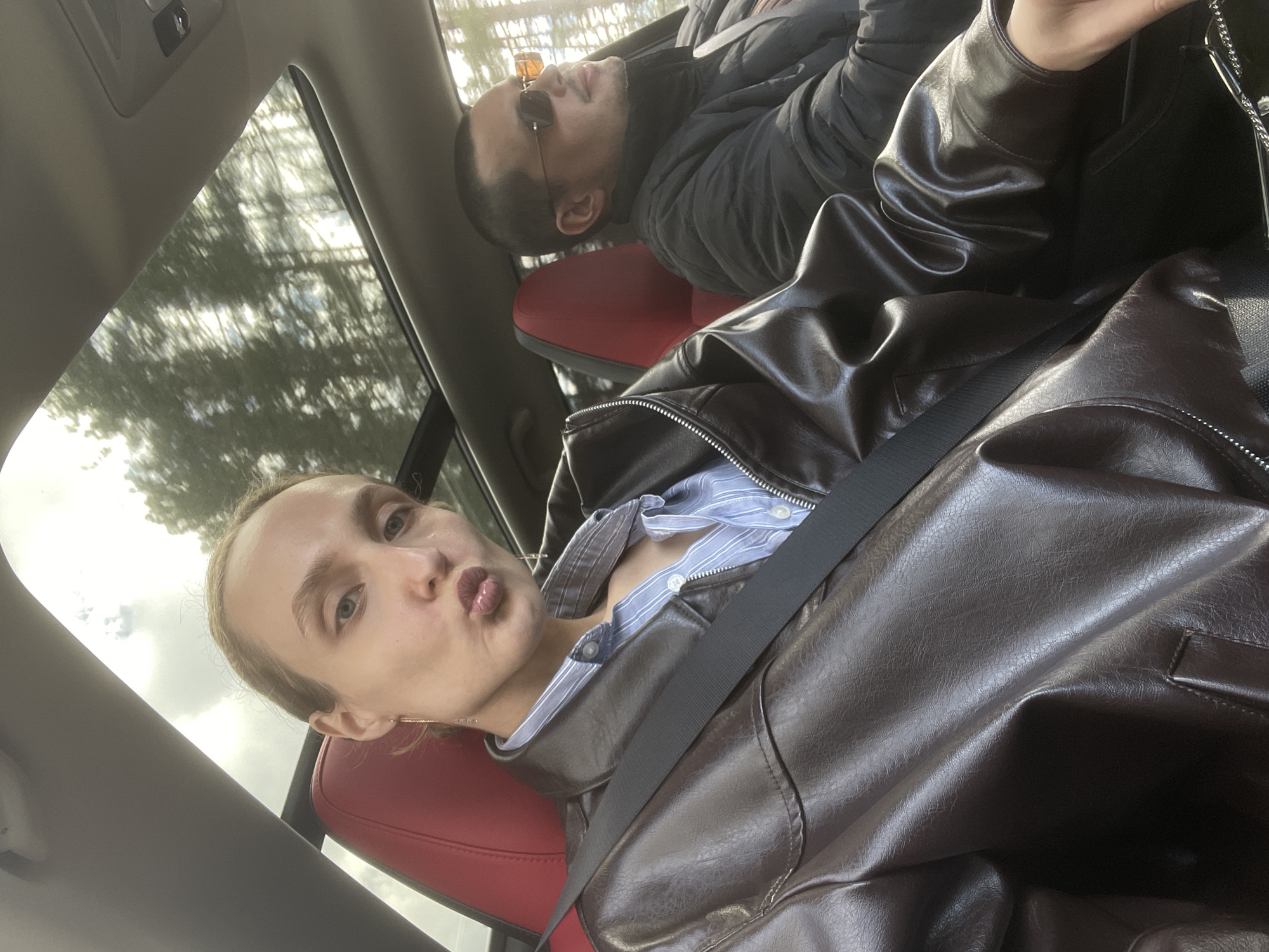 The image, rotated 90 degrees clockwise, captures two individuals inside a car, likely a selfie taken by the person in the foreground.
In the immediate foreground, a person with light skin and light hair (pulled back) looks directly at the camera with a pouting, "duck face" expression. They are wearing a dark brown leather jacket over a light blue and white striped button-up shirt, with a black seatbelt fastened across their chest. Their car seat has distinctive red accents.
In the background, another person, possibly male, is seen resting in the passenger seat. They have short dark hair, are wearing sunglasses, and are dressed in a dark puffer jacket. Their head is tilted back, suggesting they are either looking up through a sunroof or are in a relaxed, possibly sleeping, posture.
The car's interior features a gray headliner with a small light or button visible. Through the car windows, a blurred view of trees and sky is visible, indicating an outdoor setting during daytime, possibly with diffused or filtered sunlight. The car seats have a modern design with red side bolsters.
The overall scene depicts a casual moment inside a vehicle, with one person actively engaging with the camera while the other is in a relaxed state. The location context provided is Ol'khovochka, Russia. No visible text is present in the image.