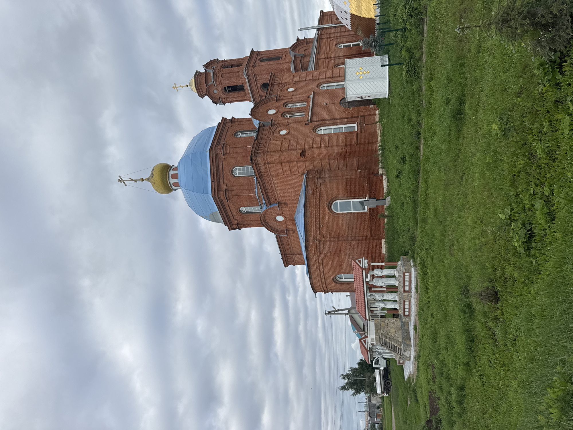 The image shows a large, red brick church with a blue dome and a smaller golden dome, located in the village of Anataş, Russia. A bell tower is attached to the main structure. In front of the church, there is a small monument with several white statues standing under a red-roofed canopy, which is accessed by a stone staircase. To the left of the monument, a white truck is parked. The church is surrounded by a green grassy area, and in the background, there are some houses and utility poles, indicating a rural or semi-rural setting. The sky is overcast with clouds.