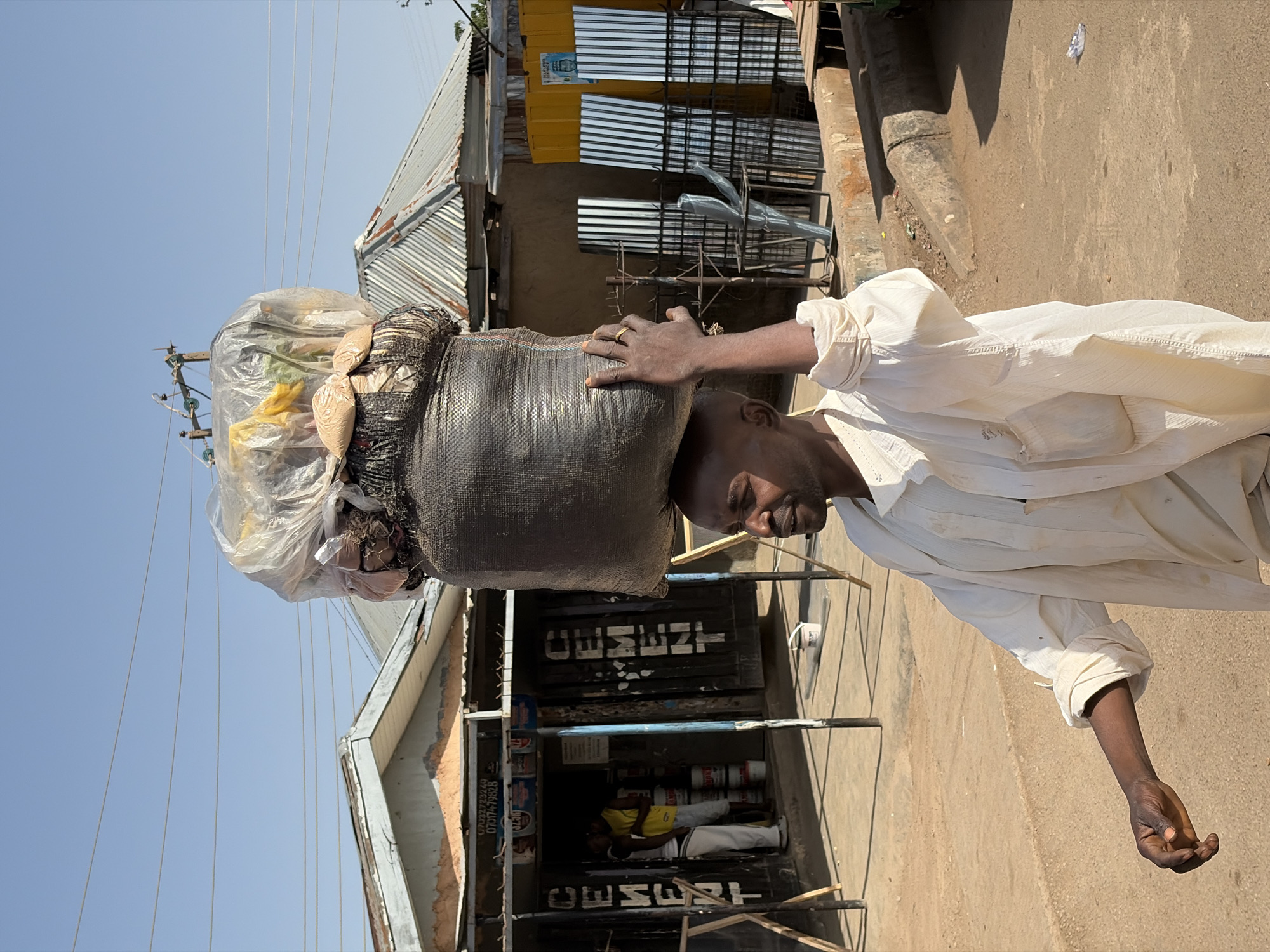 In Gura Top, Nigeria, an adult African male, facing right, balances a large, dark gray, woven bag on his head. The bag contains various items including light-colored materials and objects wrapped in clear plastic. He wears a white shirt, and his right hand rests on top of the bag. Behind him is a building with a sign that appears to read "CEMENT" vertically. Inside the building, two people are visible, one wearing a yellow shirt. Metal sheets and bars are stacked near the building. The ground is dirt-colored. The sky is light blue.
