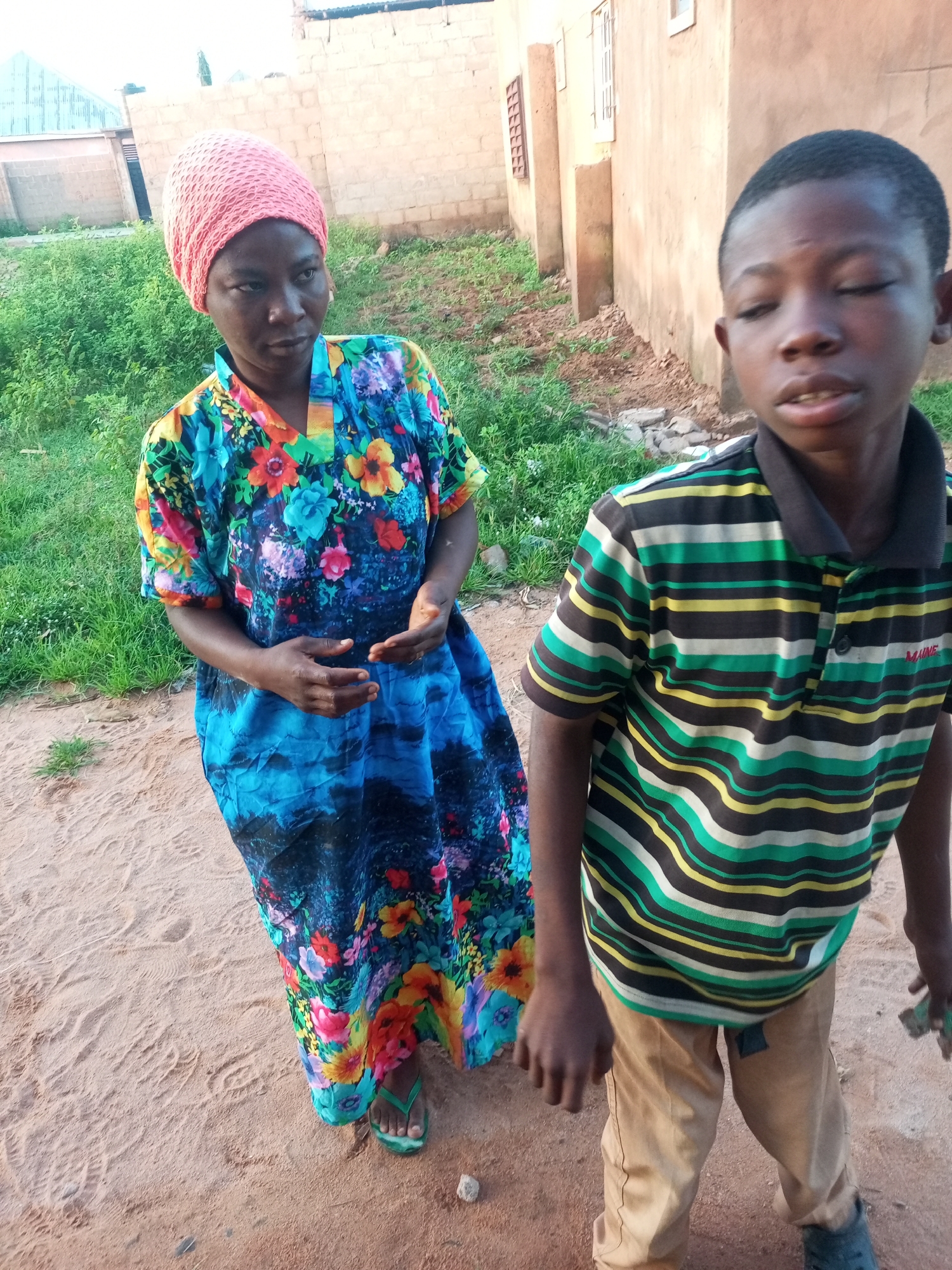 In the city of Tempure, Nigeria, a young boy and a woman stand outdoors on sandy ground. The boy, in the foreground, wears a striped polo shirt with green, yellow, black, and white bands, and khaki pants. He is looking downwards with his eyes closed. The woman, to his left, is wearing a vibrant, multicolored floral dress and a pink headscarf. Her hands are clasped in front of her. Behind them, a beige building with windows and a light-colored roof is visible, with some green vegetation growing around it. The environment appears to be a residential area.