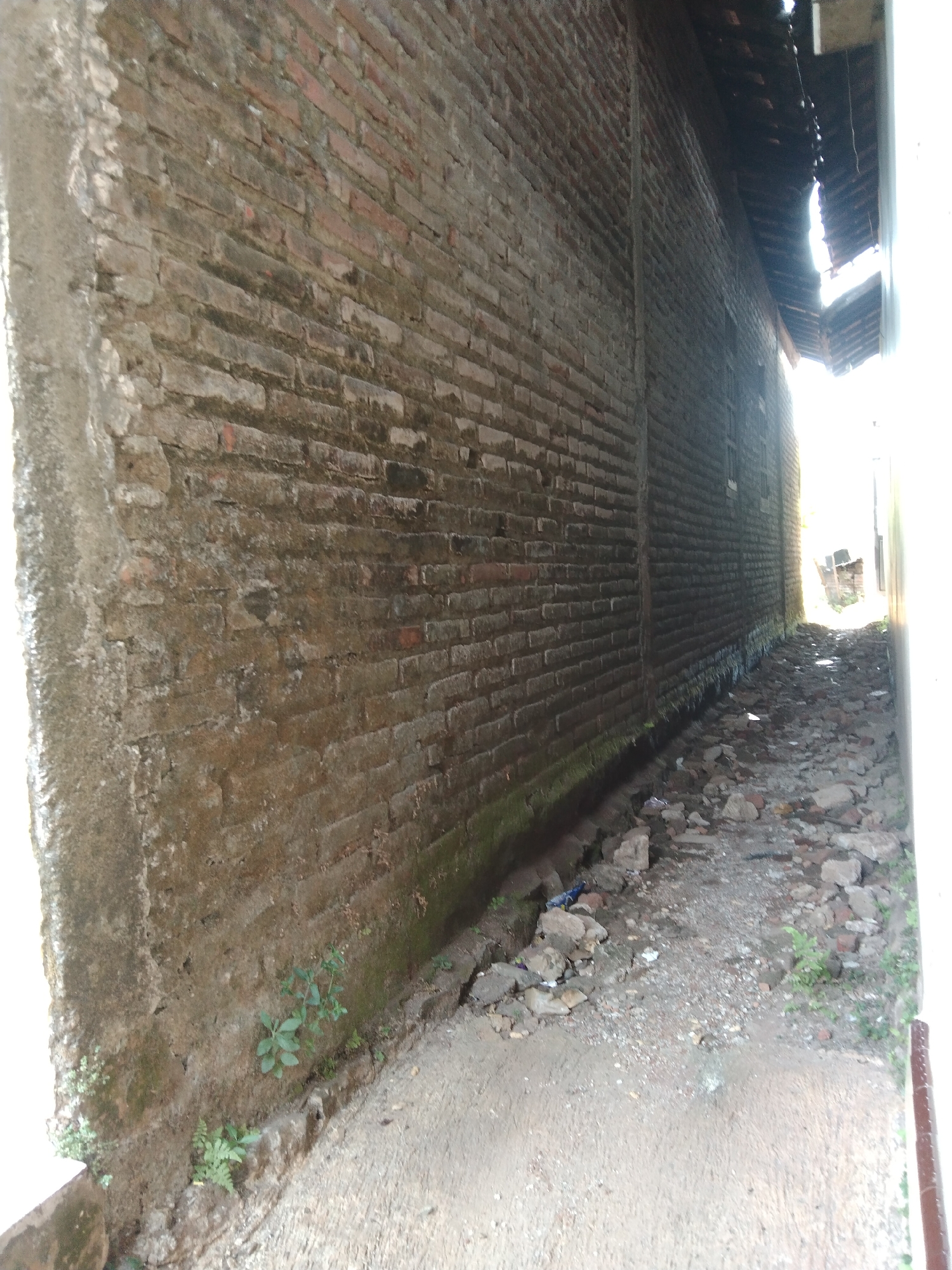 The image shows a narrow, unpaved alleyway between two brick buildings. The building on the left has a weathered, moss-covered brick wall that appears to be the exterior of a structure. The building on the right has a similar brick wall, partially obscured by the roofline of an overhang. The ground in the alley is uneven, covered with loose stones, gravel, and some scattered debris, including a blue plastic item. Small green plants are growing along the base of the left wall. The alley recedes into the distance, where sunlight is visible, suggesting an open area or the end of the alley. The overall impression is one of disrepair and neglect. The location is identified as Kelet, Indonesia.