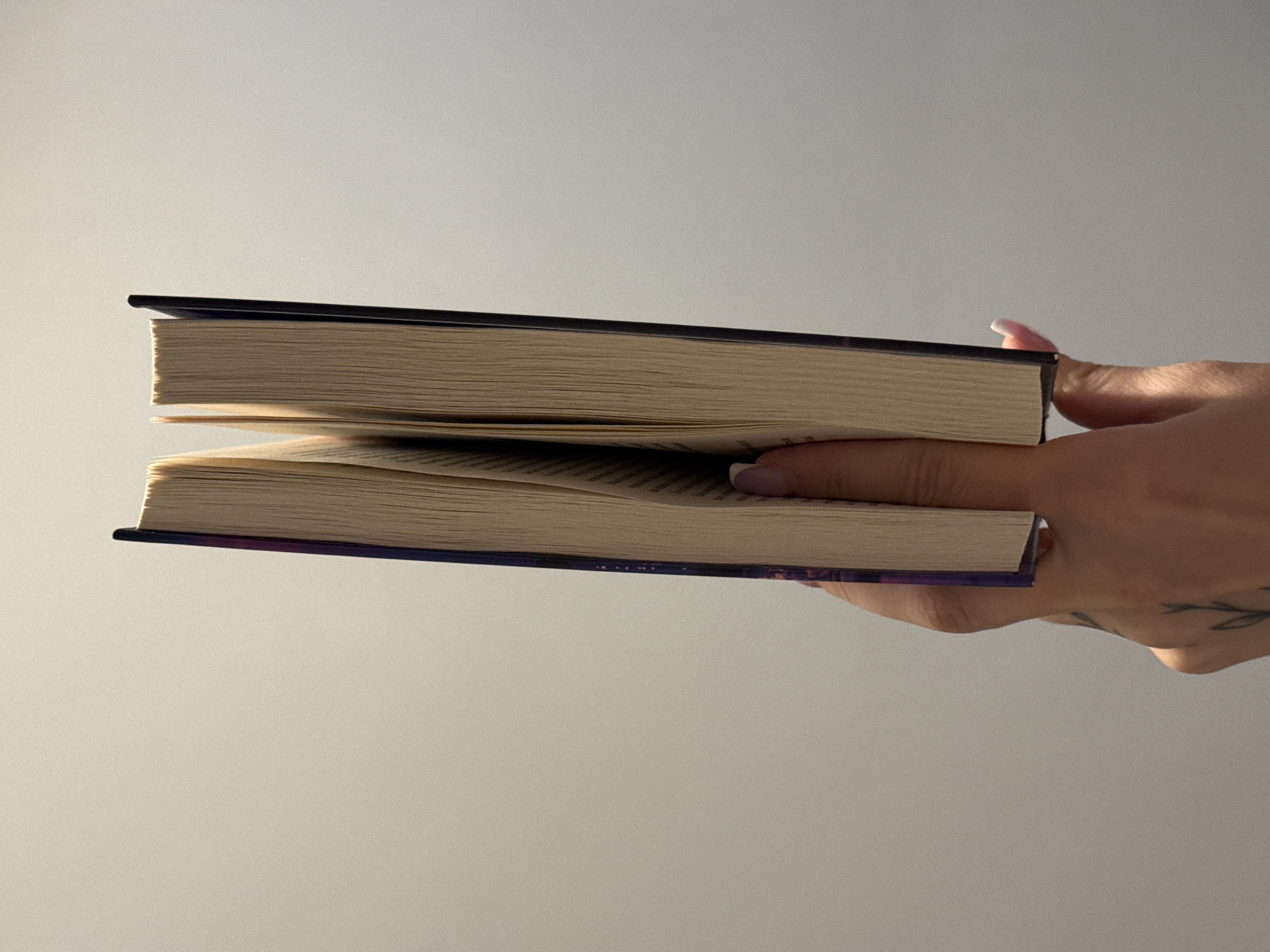 A person's hand is holding two books, one stacked on top of the other. The books are open, and the pages are visible, showing text. The top book has a dark cover, while the bottom book has a purple cover with some colorful patterns. The hand has fingernails with light pink polish and a tattoo of a plant on the wrist. The background is a plain, light gray wall, suggesting an indoor setting. The lighting appears to be soft, possibly from natural light coming from a window, as indicated by the subtle shadows. The image is taken from a slightly elevated angle, looking down at the books and hand. No specific time of day or weather conditions can be inferred. The location context of Łódź, Poland, does not provide any specific visual cues within the image itself.