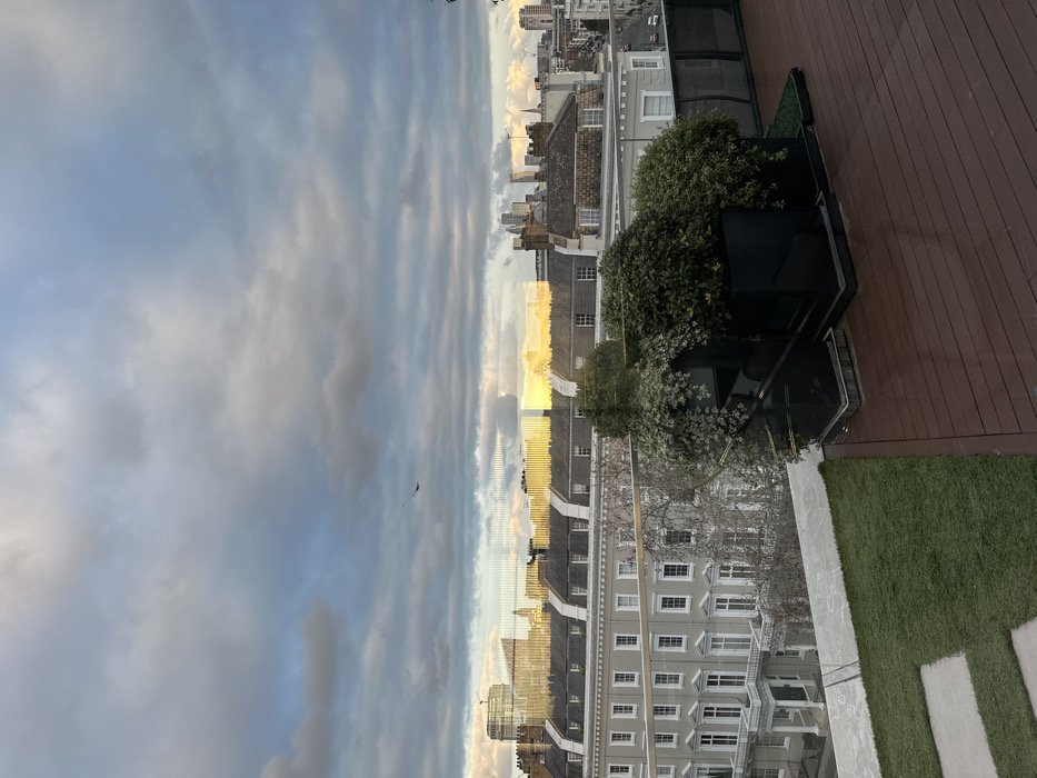 The image captures an elevated view, likely from a rooftop terrace or balcony, overlooking a cityscape. The perspective suggests a clear distinction between a built-out outdoor space in the foreground and a broader urban landscape stretching into the distance.
In the foreground, on the right side, there is a section of dark brown wooden decking. Adjacent to the decking, on the left, is an area covered with artificial green grass, characterized by a uniform, short pile. White, rectangular paving stones are partially visible embedded within this grassy area. A short glass barrier, possibly a railing, separates this terrace space from the view beyond.
Along the edge of the terrace, a series of dark, rectangular planters are visible, containing lush green foliage, including what appears to be ivy or similar climbing plants. These plants are well-established and contribute to a green, cultivated feel on the terrace.
Beyond the terrace, the midground is dominated by the rooftops and upper stories of residential buildings. These buildings feature a classic architectural style, possibly Victorian or Georgian, with numerous windows and gabled roofs. The structures appear to be made of light-colored stone or stucco. There's a prominent reflection of the sky and distant light on the windows of some buildings, suggesting they are facing the light source.
The background reveals a hazy cityscape with a silhouette of taller, more modern buildings in the far distance, indicating a larger urban center.
The sky occupies a significant portion of the upper half of the image. It is partly cloudy, with a mix of soft, streaky clouds and patches of clearer blue sky. The lighting suggests either late afternoon or early morning, with a warm, golden glow visible near the horizon line, particularly reflected in the distance and on some building surfaces. This golden light indicates the sun is low in the sky, creating long, soft shadows and highlighting the cloud formations. A small, dark bird is visible in flight against the cloudy sky in the upper left quadrant.
The overall mood is peaceful and serene, with the soft light and scattered clouds suggesting a quiet moment in an urban environment.