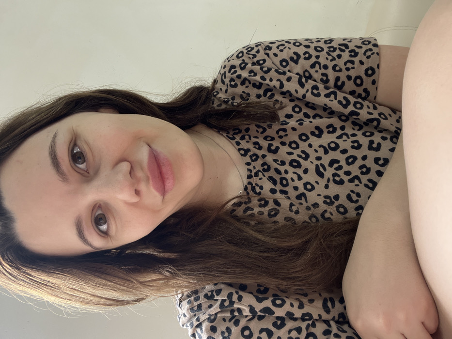 This image depicts an adult female, located indoors in the city of Leninakan, Russia. She is positioned facing the camera, making direct eye contact, with a slight upward curve to her lips. Her long, dark brown hair is parted centrally and falls over her shoulders. She wears a short-sleeved, round-neck top featuring a tan and dark brown leopard print pattern. A thin, light-colored necklace with a small circular pendant is visible around her neck. A portion of her left forearm, bent, is visible at the lower left of the frame. The background is a plain, light-colored wall, illuminated by soft, diffused lighting.