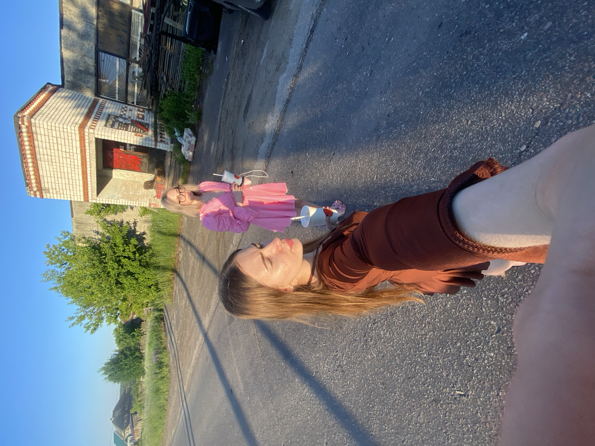 Two young women are standing on an asphalt road in front of an industrial building in Медведево, Russia. The woman in the foreground is wearing a brown dress and is holding a cup in her right hand. The woman in the background is wearing a pink dress and glasses, and is also holding a cup. The building behind them is made of white brick and appears to be abandoned or under renovation, with a red door and broken windows. The sky is clear and blue, and the sun is casting long shadows. There are trees and grass on the side of the road, and a fence can be seen in the distance.