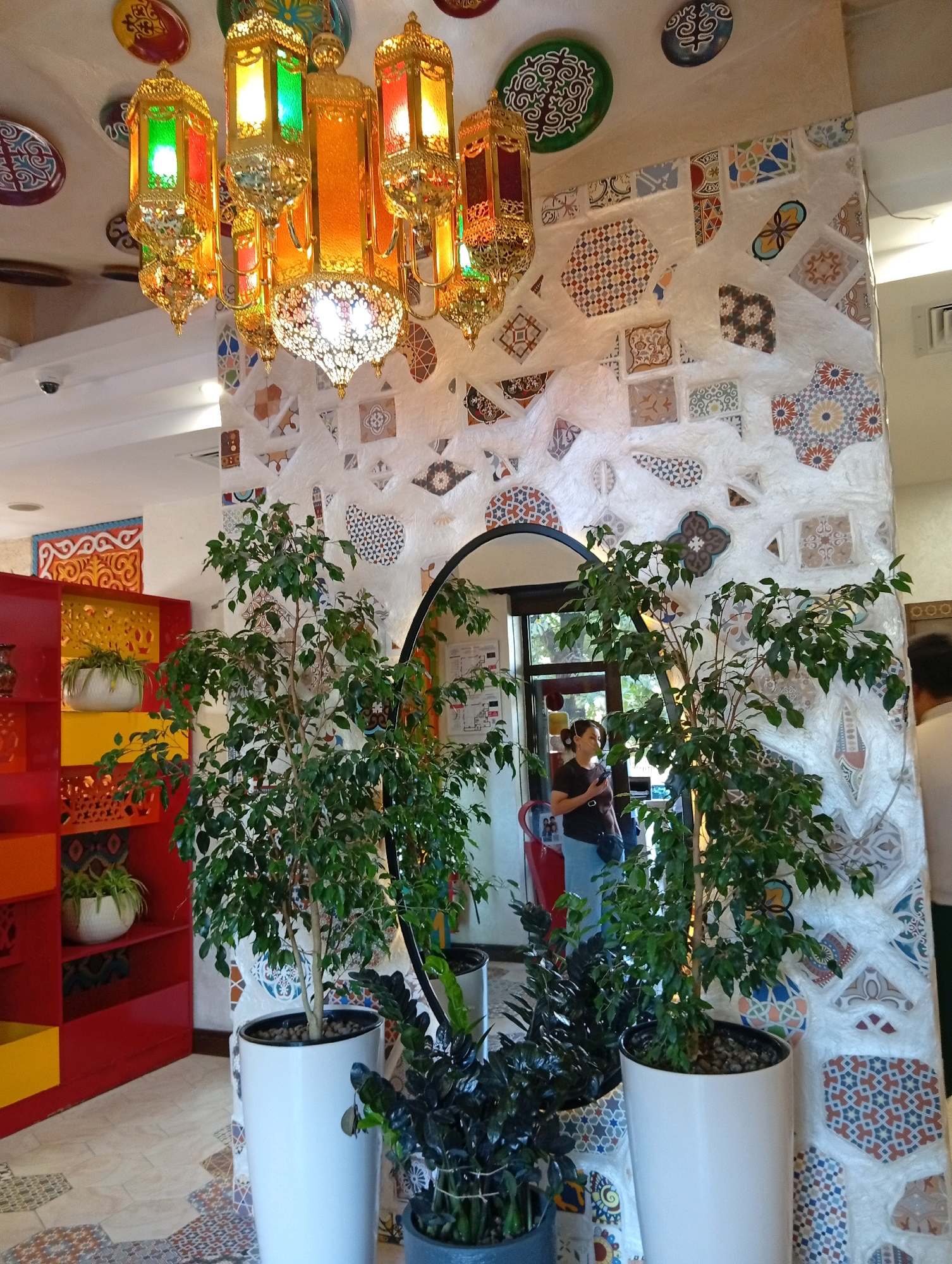 The image depicts the interior of what appears to be a restaurant or cafe in Bishkek City, Kyrgyzstan, with a distinct Middle Eastern or Moroccan theme. The dominant features are an ornate chandelier with multiple colored glass lanterns, a wall adorned with an array of colorful, patterned tiles, and two large potted plants in the foreground.
The chandelier, hanging from the ceiling, is made of gold-toned metal and features lanterns in shades of orange, red, and green. Below the chandelier, a decorative mirror with a dark frame is placed against the tiled wall.
The wall itself is a focal point, textured and embellished with a mosaic of small, irregular tiles in various geometric patterns and colors, including blues, reds, oranges, and browns. Some tiles are square, others are rhomboid or triangular, creating a dynamic and visually rich surface.
In the foreground, two large green plants in white cylindrical pots stand prominently. The plant on the right is larger and fuller, its leaves obscuring part of the mirror and the tiled wall. A smaller, darker-leaved plant is partially visible in a dark gray pot to the left of the larger plant.
To the left of the plants, a bright red shelving unit with yellow accents is visible, containing a white pot with a smaller green plant. The floor is tiled with a pattern of geometric shapes in muted colors.
A woman is partially visible in the background, standing near a doorway or window, looking towards the right. She is wearing a dark t-shirt and blue jeans and has a dark purse slung across her body. The lighting suggests it is daytime, with natural light filtering in from the background. The overall ambiance is decorative and cultural, indicative of a place aiming for a specific aesthetic.