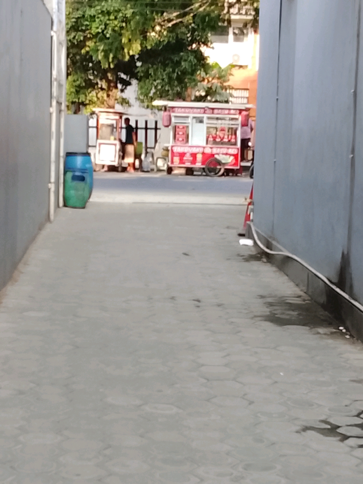 In Slawi, Indonesia, a narrow alleyway is depicted, paved with light gray hexagonal interlocking concrete blocks. On the left side of the alley, a large, blue plastic trash bin stands against a gray wall. Further down the alley, a red and white food cart is visible, with a person standing beside it. The cart appears to be selling food or drinks, as suggested by the display window and signage. Another, smaller food cart or stall is also present, with a person in a black shirt standing behind it. Trees with green foliage are visible in the background, and the buildings have orange and white facades. The scene is lit by natural daylight, casting subtle shadows on the ground.