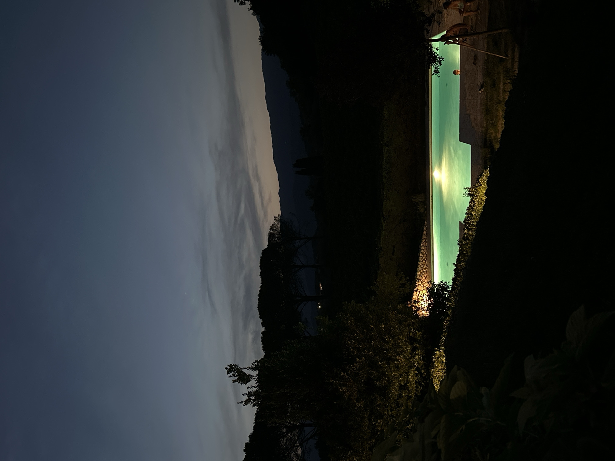 The image depicts a nighttime scene at a swimming pool, likely in a rural or secluded area, as evidenced by the silhouetted trees and hills in the background. The sky is a deep twilight blue, with faint clouds. The pool itself is illuminated from within, casting a bright, turquoise glow on the water. There are a few people visible near the pool. One person is partially submerged in the water, with their head above the surface. To the right of the pool, two people are seated on a stone wall, appearing to be relaxing. In the foreground, dark foliage and landscaping are visible, with some artificial lighting illuminating a stone wall and nearby bushes. The overall impression is one of a tranquil evening setting. The location in Conegliano, Italy, is not directly evident from the image itself, as there are no identifying landmarks.