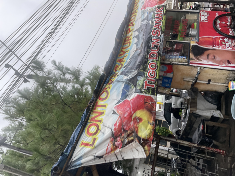 The scene is a street food stall in Ngamprah, Indonesia, with the coordinates -6.867 latitude and 107.517 longitude. The stall is covered by a large, colorful banner that reads "Lonton Sayur Padang" and "Tigo Dun Sanak." The banner features a red flower and a building. To the right of the stall is a silver car and a person sitting in the car. To the left of the stall is a metal gate and a black object. On the stall, an advertisement for the beverage "Puc** Har**n" is displayed. There are numerous power lines stretching above the stall. The sky is grey and overcast. The scene is wet, likely due to rain.