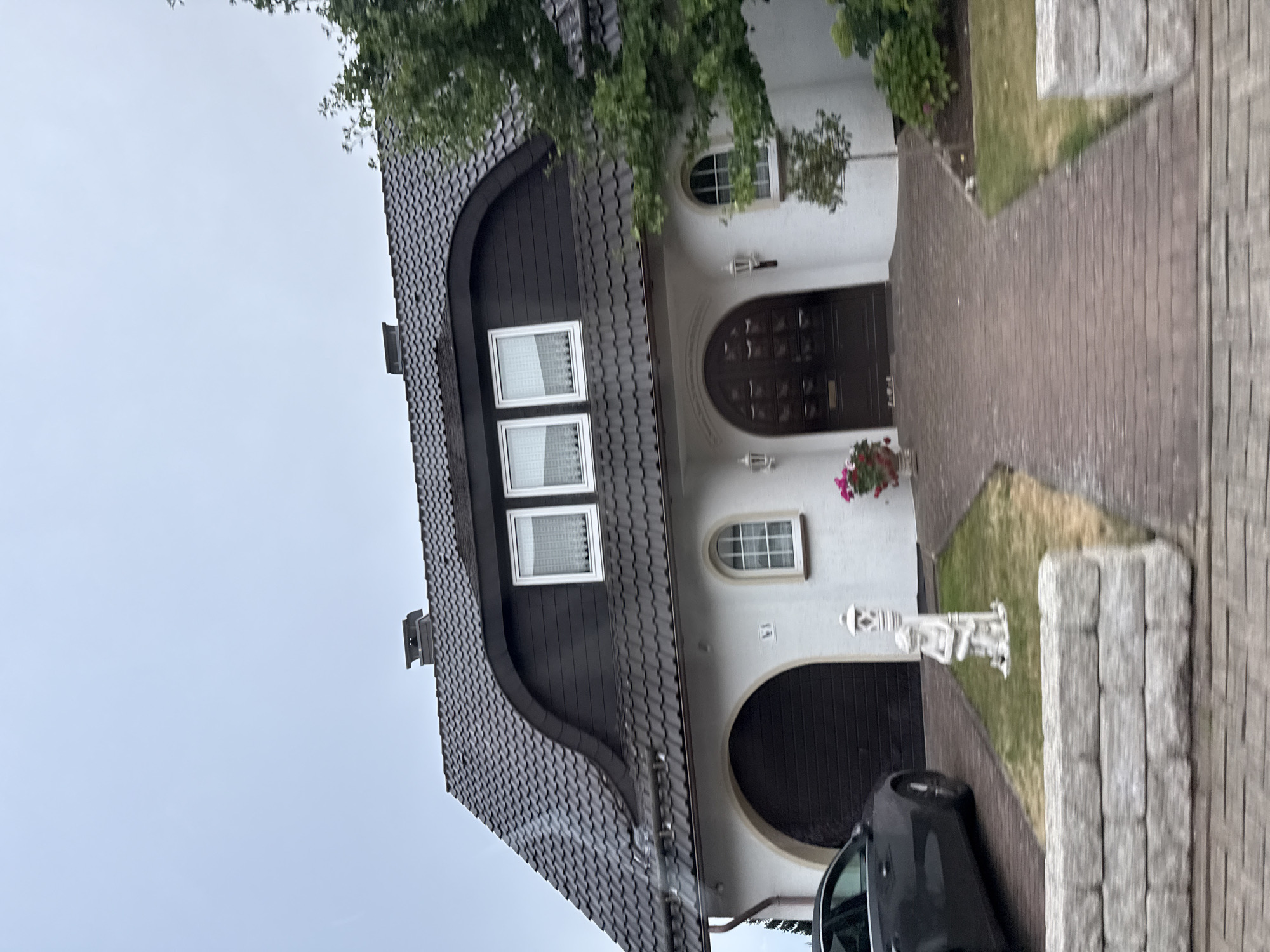 The image shows a detached house with a dark, tiled, mansard roof. The house has a white facade with a large, arched, dark brown garage door on the left side and a similarly arched, dark brown double-door entrance on the right. Above the entrance are two white-framed windows with white curtains. To the left of the entrance, there is a smaller arched window with a white frame. In front of the garage, a dark gray car is parked. A paved driveway leads to the entrance, bordered by grass and some greenery. A white decorative statue of a child holding a lamp post stands on the grass to the left of the driveway. A low, grey stone wall is visible in the foreground. A tree with green foliage is on the right side of the house. The sky is overcast. The house is located at Marktstraße 61 in Bad Sachsa, Germany.