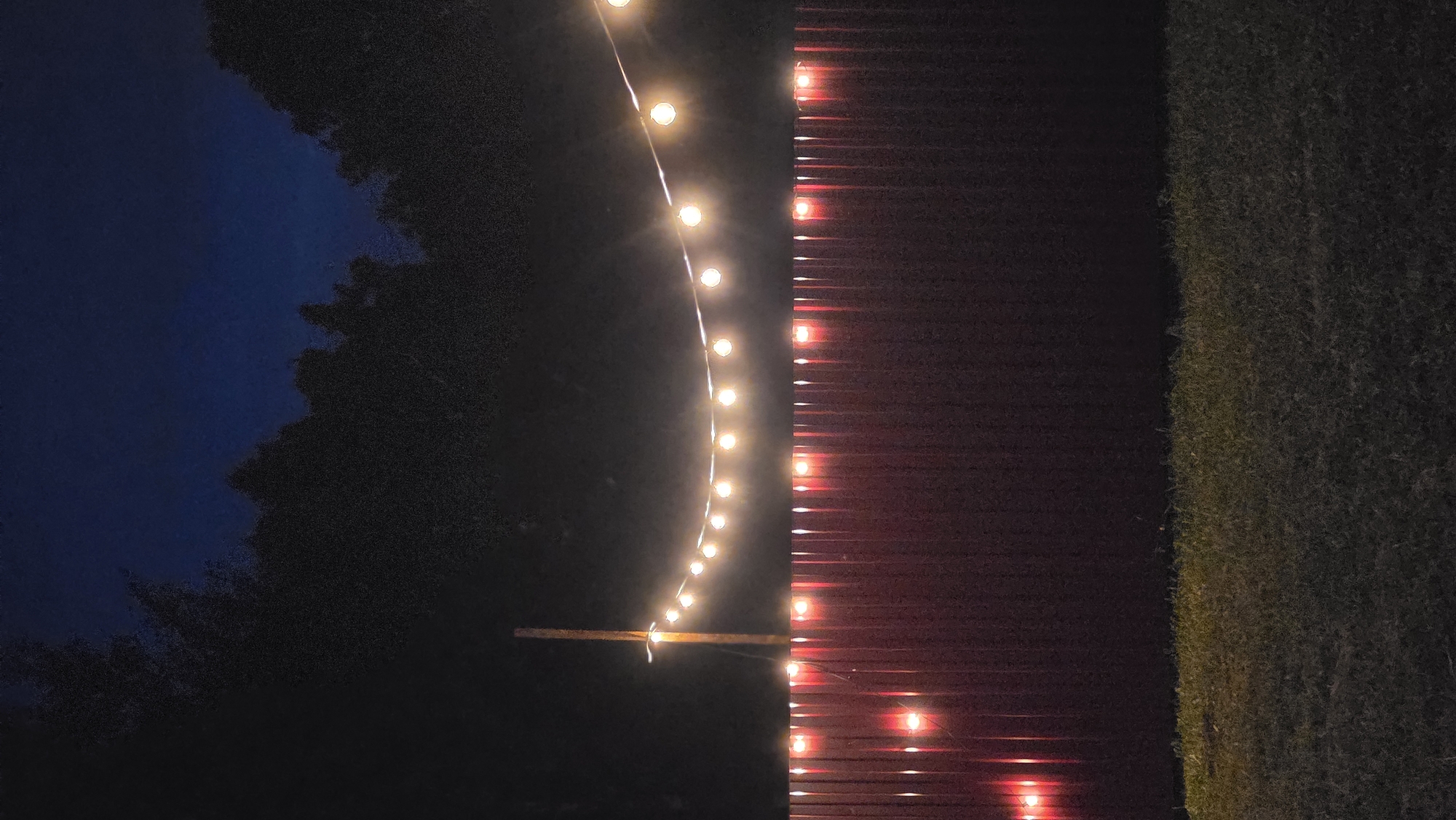 The image depicts an outdoor scene at night. A string of warm-toned lights is strung across the frame, supported by a wooden post on the left. Below the lights is a tall, dark red fence with vertical corrugated panels. Some of the fence panels are illuminated from behind by red lights, creating a glow. The ground in front of the fence is covered in grass, which is dimly lit. In the background, silhouetted trees are visible against a dark blue night sky. The overall atmosphere is serene and the image appears to be taken from an unknown outdoor location.