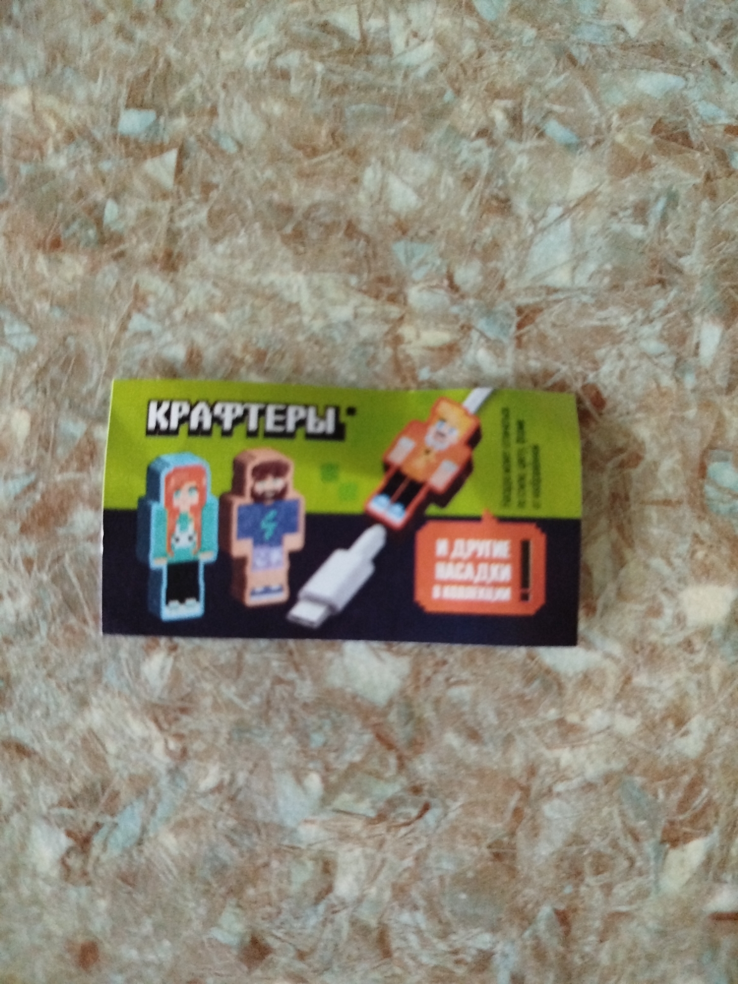 The image shows a small, rectangular, green and black package on a textured, mottled surface that appears to be a countertop or table. The package features pixelated graphics of characters that resemble those from the video game Minecraft. The text on the package is in Cyrillic. The phrase "КРАФТЕРЫ" is prominently displayed in white block letters on the green portion. Below this, two figures are depicted: one in a blue hooded outfit with red hair and the other in a purple outfit with a beard. To the right, a white cable-like object is shown connecting to an orange and yellow pixelated figure. In the lower right corner, an orange rectangular icon contains Cyrillic text that translates to "and other attachments". The overall presentation suggests a toy or novelty item related to the Minecraft franchise. The background surface has a swirling, fibrous pattern in shades of brown, tan, and light blue. The image appears to be taken from a slightly overhead angle.