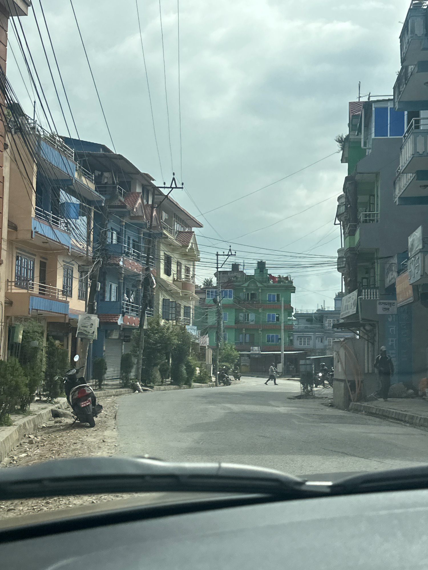 The image depicts a street scene in Kageshwori Manohara, Nepal, likely taken from within a vehicle. The street is paved, with a slight curve to the right. On the left side of the street are multi-story buildings with balconies and visible electrical wiring crisscrossing overhead. A black motorcycle is parked on the left side of the road, near the curb. Further down the street, more buildings and some trees are visible. On the right side of the street, there are also multi-story buildings with balconies and what appear to be shops or businesses at ground level. A few individuals are present on the street, including one person walking in the middle of the road and another standing on the right sidewalk. The sky is overcast.