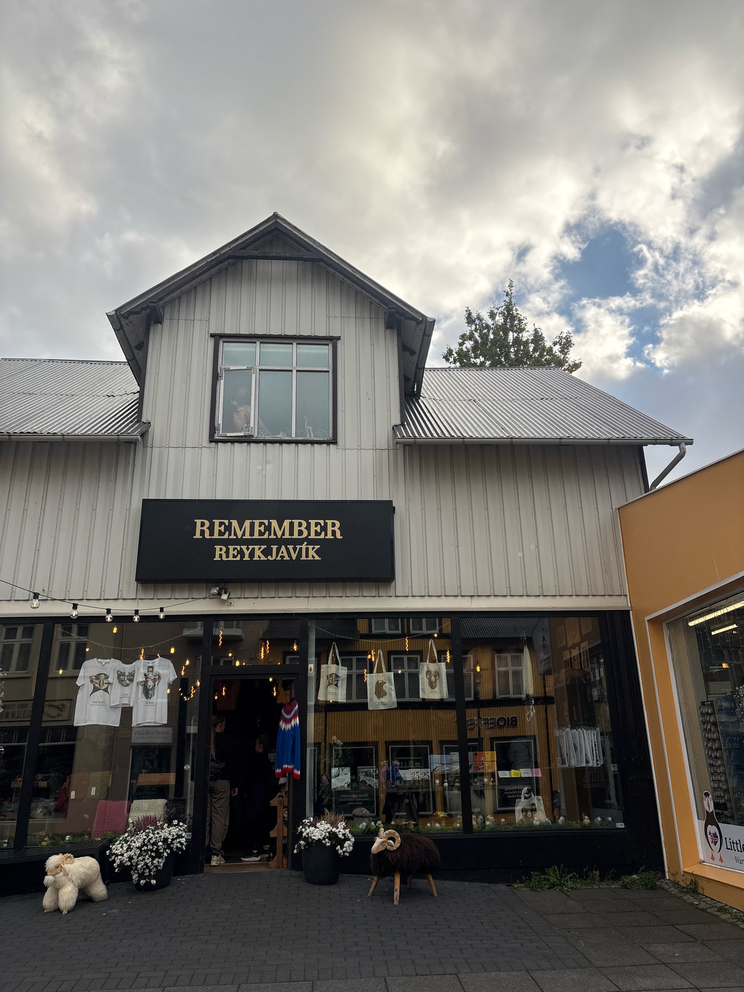 The image shows the exterior of a store named "REMEMBER REYKJAVÍK" located in Reykjavik, Iceland. The store has a white, corrugated metal facade with a prominent black sign above the entrance. The entrance features large glass windows displaying merchandise, including t-shirts and tote bags. Outside the store, there are decorative elements such as strings of lights, flower pots, and two sheep-like sculptures. A tree is visible behind the store, and the sky is cloudy. Adjacent to the store on the right is another building with an orange facade. The ground is paved with dark bricks. The scene appears to be a street-level view of a commercial establishment.