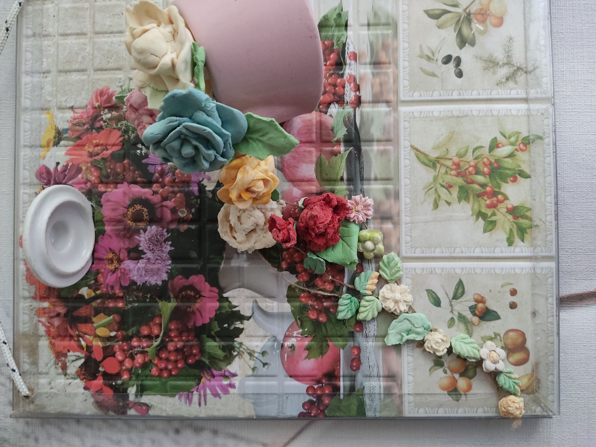 The image depicts a decorative plaque, possibly a wall hanging, featuring a collage of floral imagery and small, three-dimensional floral embellishments. The plaque is displayed against a textured, light-colored surface. At the top of the plaque, a white lid or cap is attached. The main visual element is a printed image of a vibrant bouquet of flowers, including pink, red, and yellow blossoms, interspersed with red berries. Superimposed on this printed image are several realistic-looking floral elements crafted from a pliable material, such as clay or fondant. These include a large blue rose, a pale yellow rose, and a cream-colored flower. Red and green leaves are also present, integrated into the design. The bottom portion of the plaque features a repeating pattern of tiled images, each depicting a branch with fruits or olives. Small, three-dimensional flowers and leaves in pastel shades of cream, beige, and green are arranged in a vine-like pattern across the lower section of the plaque. The plaque is edged with a clear plastic material, and a white cord with blue accents is attached to the top for hanging. The setting provides no specific information about the city of Noginsk, Russia, or any urban context.