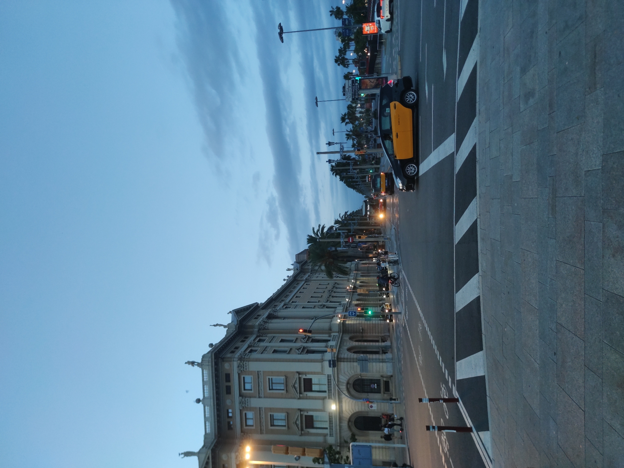 The image depicts a wide street in Barcelona, Spain, at dusk. A grand, ornate building with classical architectural elements lines the left side of the street, featuring multiple stories, arched entrances, and statues adorning its roofline. Traffic lights are visible, with one showing red and another green, indicating an intersection. Several vehicles are present, including a yellow and black taxi in the foreground, a white car, and a bus further down the street. Pedestrians are seen on the sidewalks and crossing the street, some on electric scooters. Palm trees are scattered along the sidewalks, adding a tropical touch to the urban landscape. The sky is a deep blue with streaks of clouds, suggesting the transition from day to night. The street is paved with asphalt, and a pedestrian crossing with white stripes is prominent in the foreground. The overall scene conveys a sense of city life in motion as evening approaches.