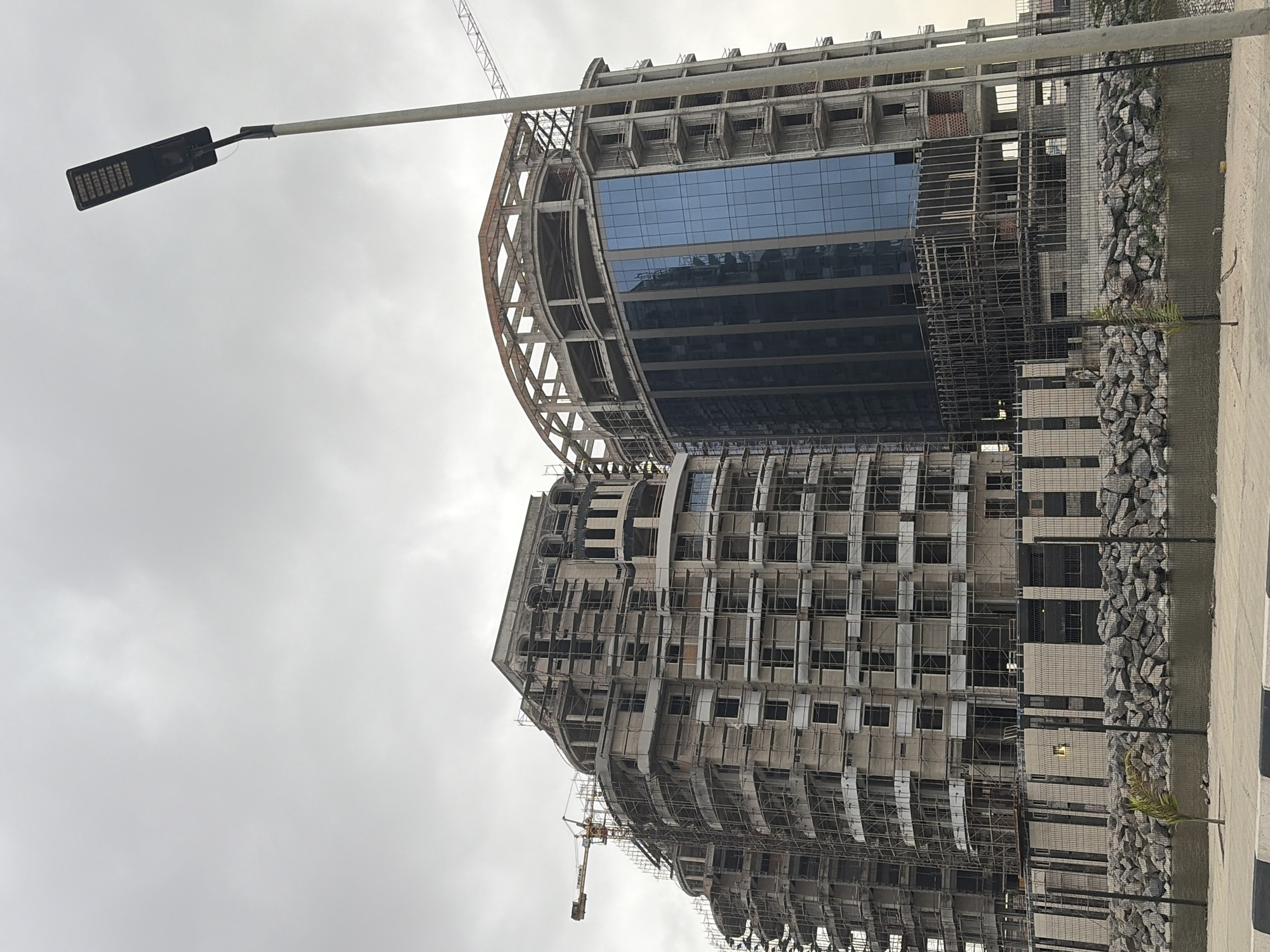 The image shows a cloudy sky over a construction site in Ilado, Nigeria. Two large buildings are under construction, both with curved facades. The building on the left is covered in scaffolding, with a crane visible above it. The building on the right has a glass exterior on its lower floors, with exposed concrete and scaffolding on the upper levels. A street light stands tall on the right side of the frame. In the foreground, a rocky embankment lines the edge of a body of water, with a black metal fence separating it from a paved area. A small tree is planted near the water's edge.