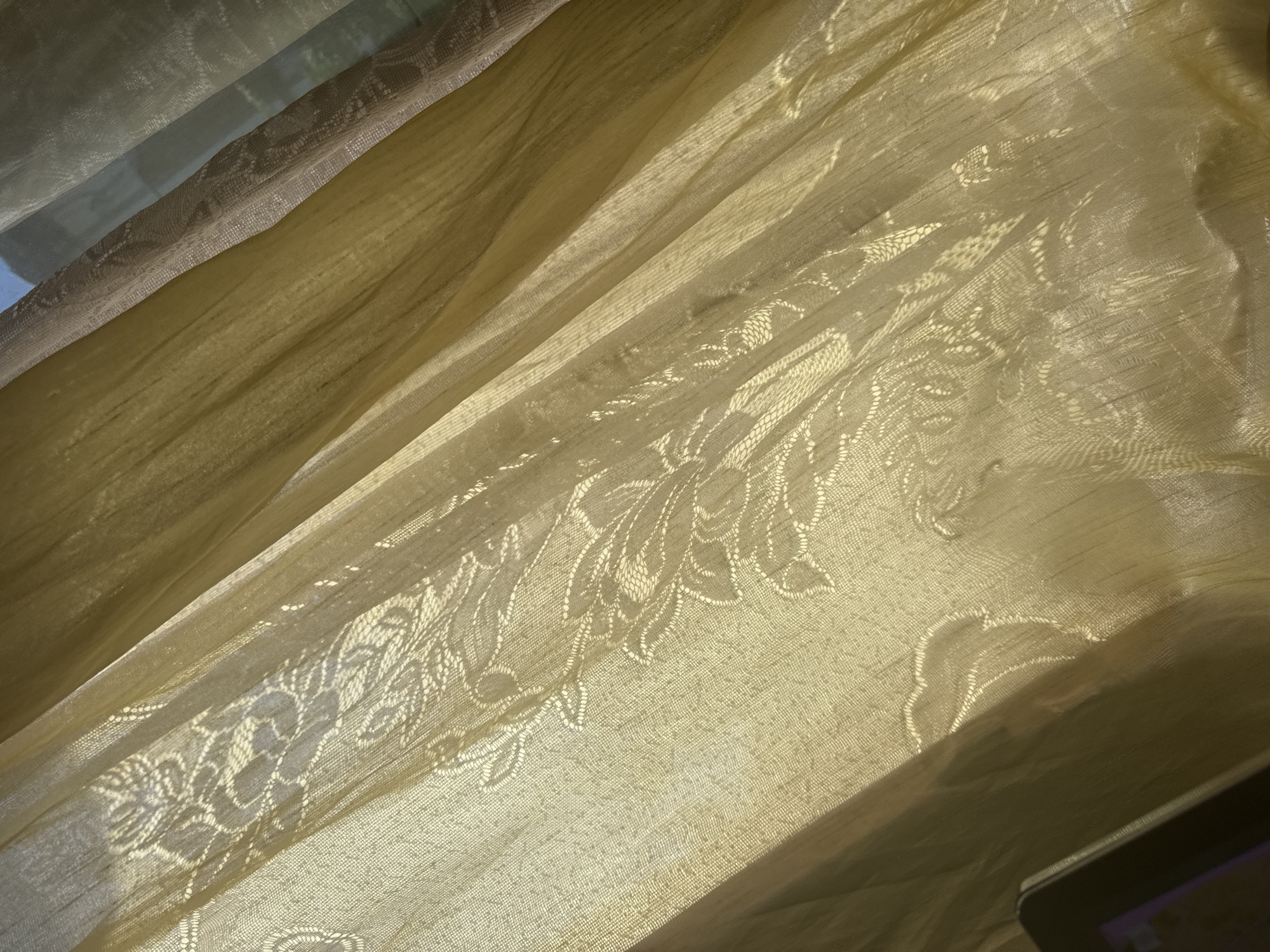 The image shows a close-up of sheer, golden curtains with a floral lace pattern. The sunlight is filtering through the fabric, creating shadows and highlighting the texture of the material. The curtains are draped, with visible folds and pleats, suggesting they are hanging in a window. In the lower left corner of the image, a dark object, possibly a tablet or e-reader, is partially visible. The scene is likely indoors, in a domestic setting, in the city of Bezden, Bulgaria.