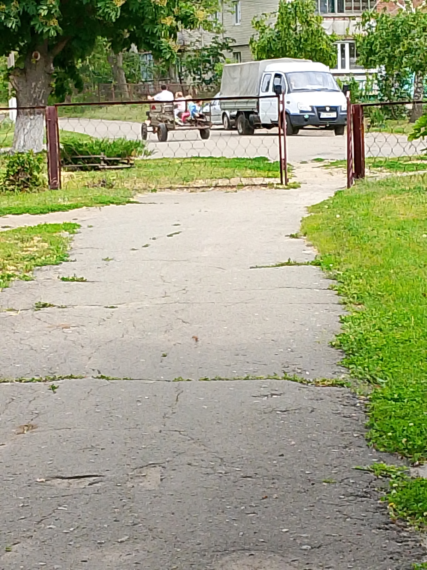 The image depicts an outdoor scene featuring a paved pathway leading towards a street or open area, viewed through a brown metal fence. In the mid-ground, a person wearing a white shirt is driving a small, open-air motorized vehicle, resembling a homemade tractor or cart. Behind this vehicle, a trailer carries several children. Further back, a white utility van with a canvas-covered bed, similar to a GAZelle model, is parked or moving slowly.
The pathway in the foreground is made of cracked asphalt or concrete, with numerous small weeds and grass growing through the fissures. Lush green grass and foliage flank the path and extend beyond the fence. In the background, beyond the vehicles, dense green trees and bushes are visible, along with parts of residential buildings, possibly apartments. The fence, with its wire mesh and sturdy posts, extends across the width of the image at eye level.
The activity involves the person driving the small vehicle with children in tow, suggesting a leisurely ride or local transport. The scene occurs during daylight hours, likely under bright, fair weather conditions. The presence of the specific utility van model and the general environment hint at a possible Eastern European or post-Soviet location. A license plate is visible on the front of the white van, but the characters are unreadable due to blurriness.