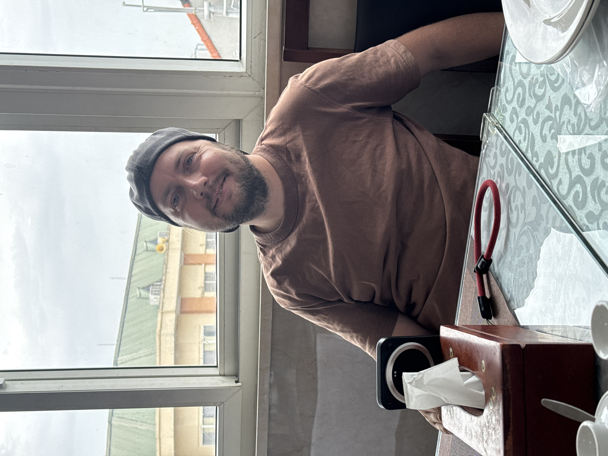 A man wearing a gray baseball cap and a brown t-shirt is seated at a table indoors, facing the camera. He has a beard and is smiling. In front of him on the table is a wooden tissue box with a white tissue sticking out, a black circular object, and a red cord. The table has a glass top with a patterned design. Behind the man is a window with a view of buildings and a cloudy sky. The overall setting appears to be a restaurant or cafe in Hanoi, Vietnam.