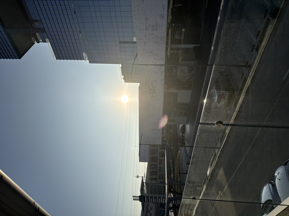 The image depicts an urban landscape under a clear, pale blue sky with a low, intensely bright sun positioned slightly right of center, casting strong light and a lens flare. The scene is viewed from an elevated position behind a clear glass balustrade with metal fasteners. An angular, grey structural element partially obscures the upper left corner.
In the mid-ground, a broad asphalt surface, marked with white lines indicating parking spaces or lanes, stretches across the scene. Several vehicles are present: parts of two silver/grey cars are visible in the bottom-left foreground, and a dark grey SUV is positioned in the mid-ground asphalt area, reflecting sunlight. Another dark vehicle is visible further back. A tall, black street light pole stands in the mid-ground, slightly right of center.
Dominating the right side of the frame is a modern, multi-story glass skyscraper with a dark framework, reflecting the sky. Adjacent to it, in the center-right, is a large commercial building featuring a light-colored facade with an irregular pattern of circular perforations. The letters "DS" and "BS" are visible on this perforated facade. Below this, darkened storefronts are present. To the left of this central building, an elevated, grey, angular structure, possibly a pedestrian bridge or ramp, is visible, partially obscuring additional commercial buildings in the background. Partial text, resembling "DOMTELAS," is discernible on one of these background buildings. Several indistinct human figures are visible on the ground level, near the commercial buildings.
Based on the architectural features, including the perforated facade, modern glass towers, and visible branding elements, the location is identified as the **Angelópolis Lifestyle Center area in Puebla City, Mexico**.