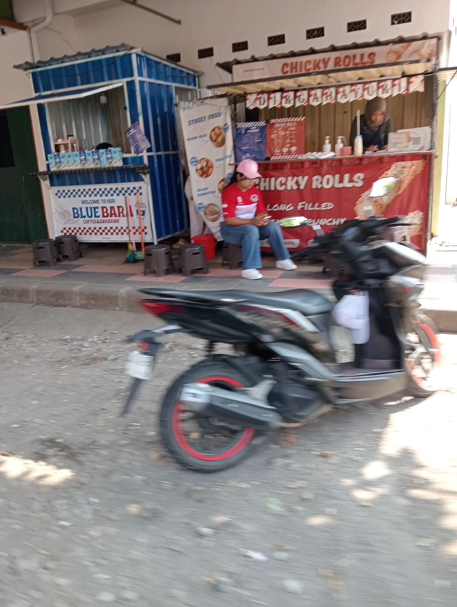 This image shows a street food stall in Grabag, Indonesia. On the left, a blue and white food stall displays beverages in cartons and has a sign that reads "WELCOME TO OUR BLUE BAR COFFEE & BARAKAN". In the center, a person wearing a red shirt, blue jeans, and a pink cap sits on a small stool, looking down at their phone. To the right of this, a stall with a red banner advertises "CHICKY ROLLS" and displays images of food. A woman wearing a hijab and an apron stands behind this stall, appearing to be serving customers. In the foreground, a black motorcycle is parked, with its wheels and part of its body blurred due to motion. The overall environment is an outdoor street setting with a paved walkway.