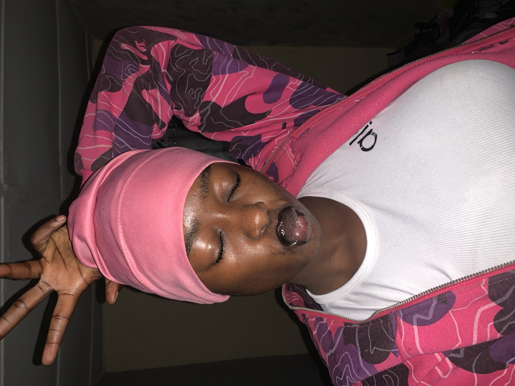The image shows a dark-skinned individual wearing a pink head covering and a pink and purple camouflage jacket over a white t-shirt. The person is lying down with their eyes closed and mouth slightly open, tongue exposed. Their hand is outstretched towards the left. The background is dark, with indistinct walls. No specific location details are discernible to indicate it's Rafin Par, Nigeria.