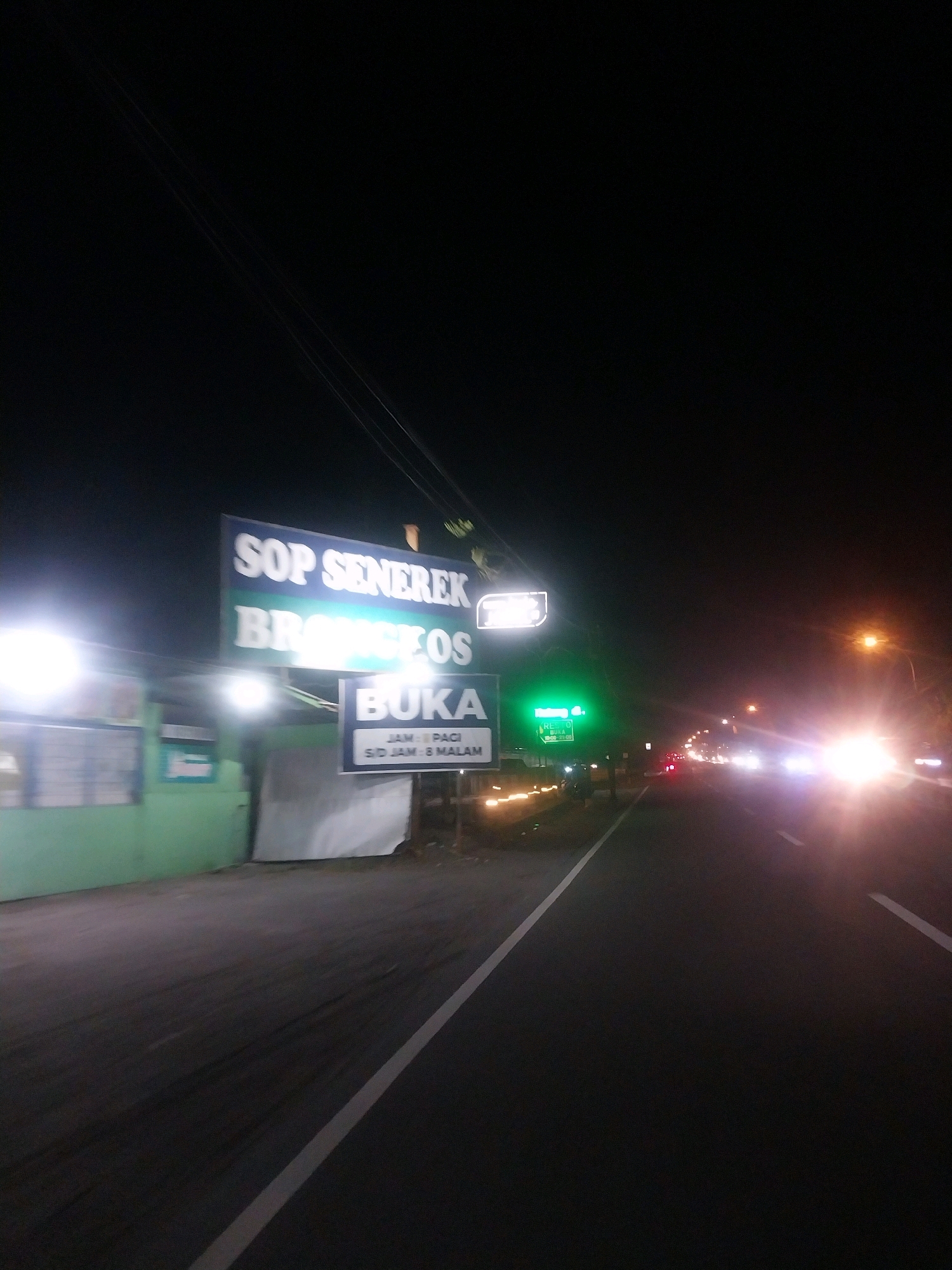 This is a night-time, eye-level, slightly blurred photograph of a road in Tempel, Indonesia. A dark, asphalt road stretches into the distance, marked with a white line on the right side. To the left of the road, a light green building with white-barred windows is visible. Above the building, a large, illuminated sign reads "SOP SENEREK BRONZEOS" in blue and white text. Below this, a smaller sign with a white background and blue text states "BUKA" and beneath it, "JAM : PAGI S/D JAM : 8 MALAM" indicating opening hours. Further down the road, a green illuminated sign is visible, and further still, headlights from approaching vehicles illuminate the scene. The sky is dark, with a few wires visible overhead.