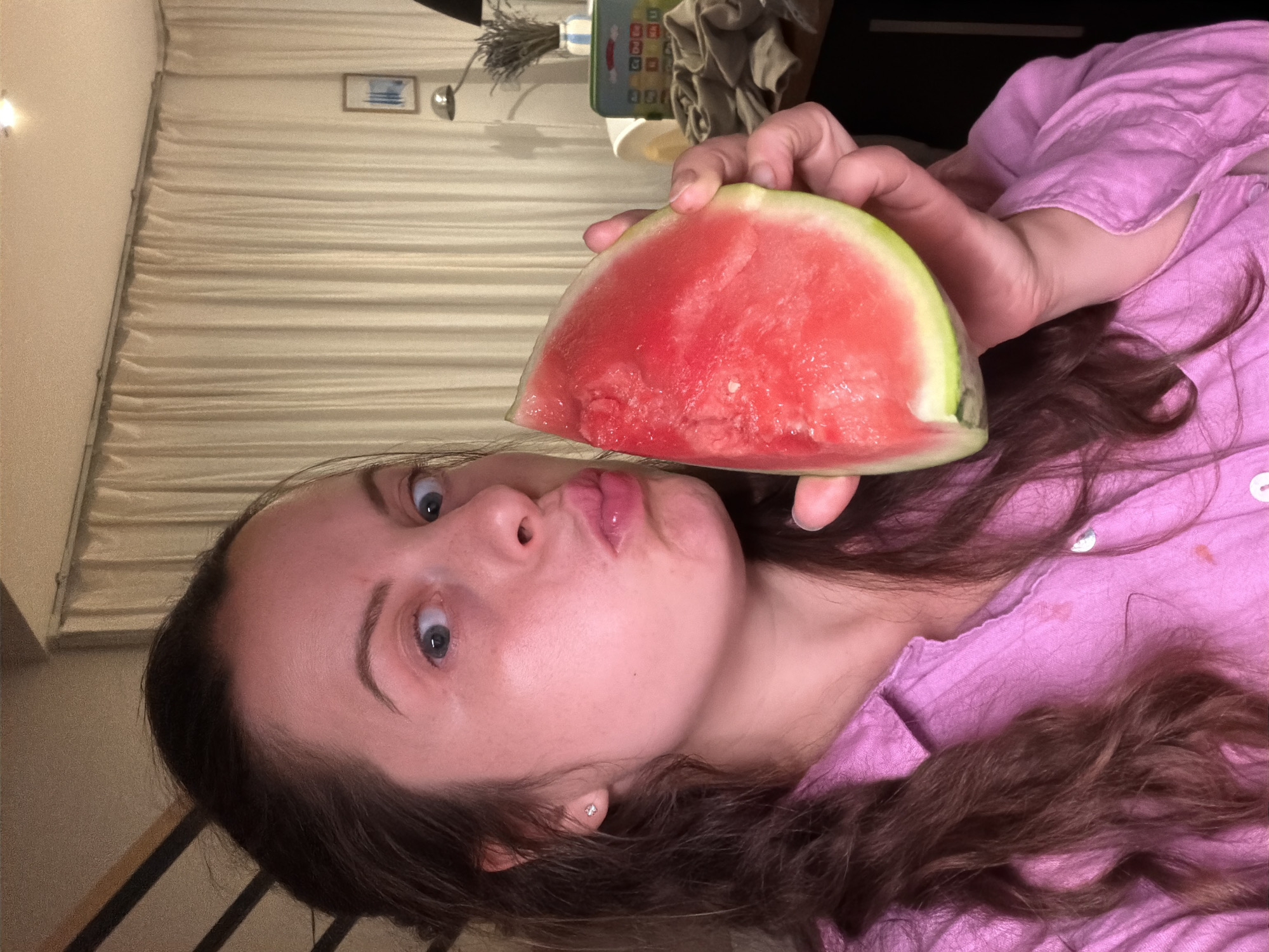 The image shows a woman holding a slice of watermelon to her face, with her lips pursed. She is wearing a pink shirt. The background includes curtains, a lamp, and a shelf with some items on it. There is no information in the image to indicate that it is located in Gangloff, Germany.