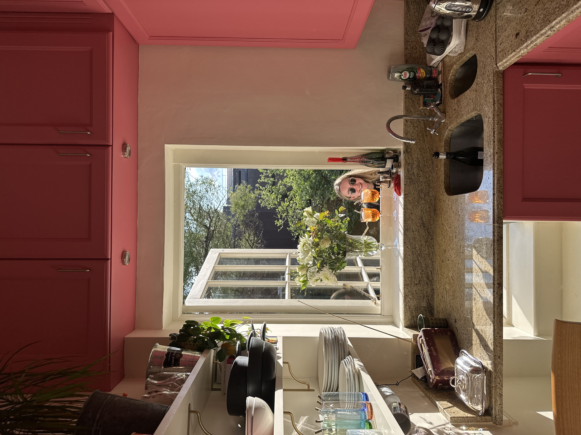 A person wearing sunglasses is looking into the kitchen of a house in London, United Kingdom, through an open window. The kitchen has pink cabinets, a granite countertop, and a white shelving unit. The shelving unit holds pots, pans, plates, and glassware. A sink is to the right of the window. Outside the window are trees and buildings. Two glasses of amber liquid and a vase of white flowers sit on the window sill.