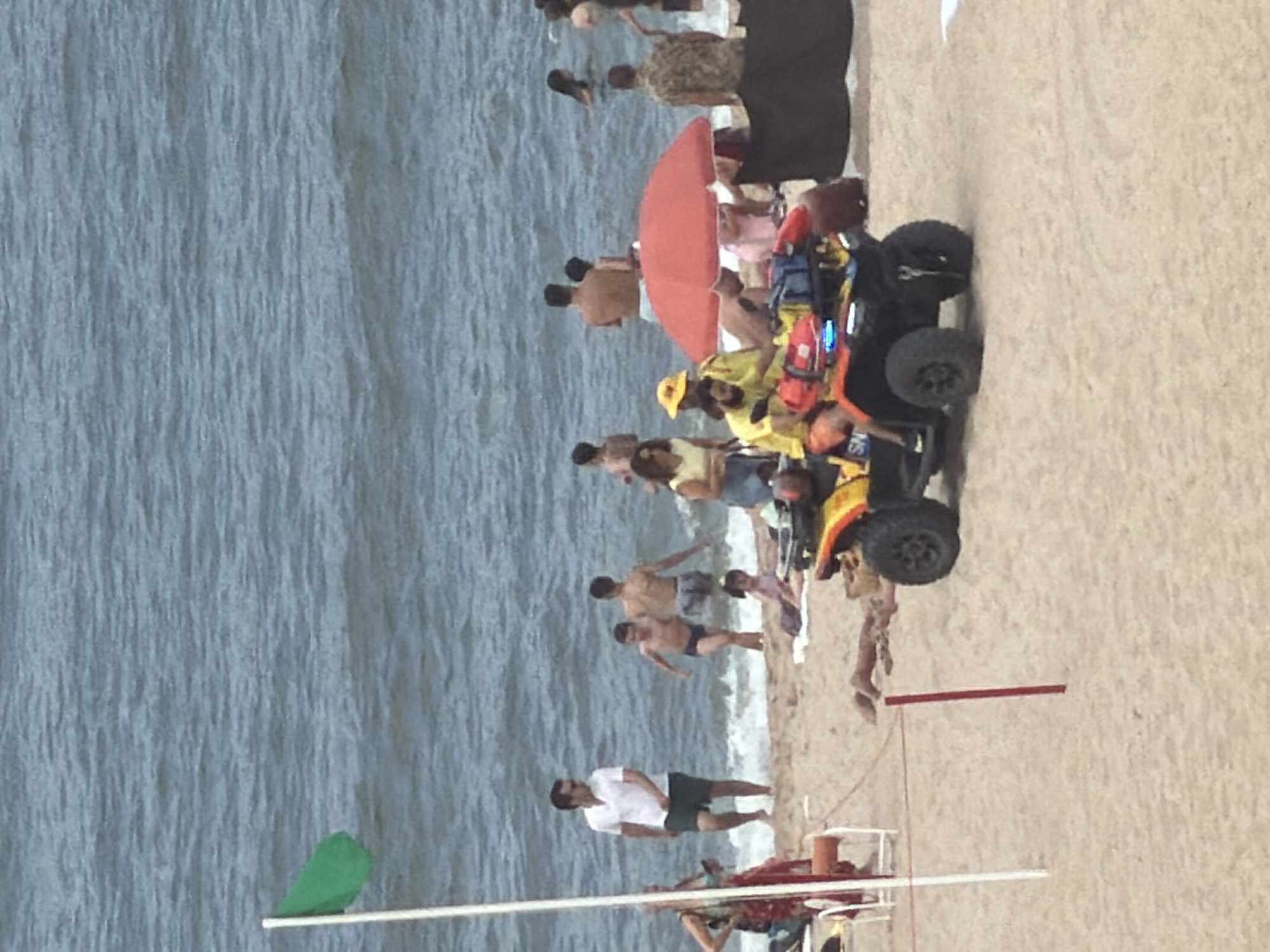 This image depicts a sunny day at a beach, likely in Espinho, Portugal, given the presence of beachgoers and the general coastal environment. In the foreground, an all-terrain vehicle (ATV) with a lifeguard crew is positioned on the sandy shore. The ATV is equipped with emergency lights and rescue gear, suggesting its readiness for an incident. Two lifeguards, one in a yellow shirt and the other in an orange top, are visible on or near the ATV.
Further back, several individuals are enjoying the ocean and the beach. Some are wading in the shallow water, while others are relaxing on the sand. A green flag is planted near the water's edge, indicating the current beach conditions. The ocean appears calm with gentle waves. The background shows more people scattered across the beach and in the water, with beach umbrellas providing shade. The overall atmosphere is one of leisure and safety under the supervision of lifeguards.