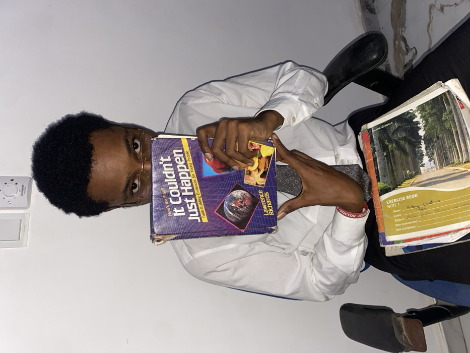 A young man in a white shirt and tie holds a book titled "It Couldn't Just Happen" by Lawrence Richards. The book's cover features a globe and colorful images. Additional exercise books are visible, one labeled "EXERCISE BOOK NOTE 1." The man wears glasses and a wristband with text on it. He sits in a chair with a black back, positioned against a white wall with a visible switch.