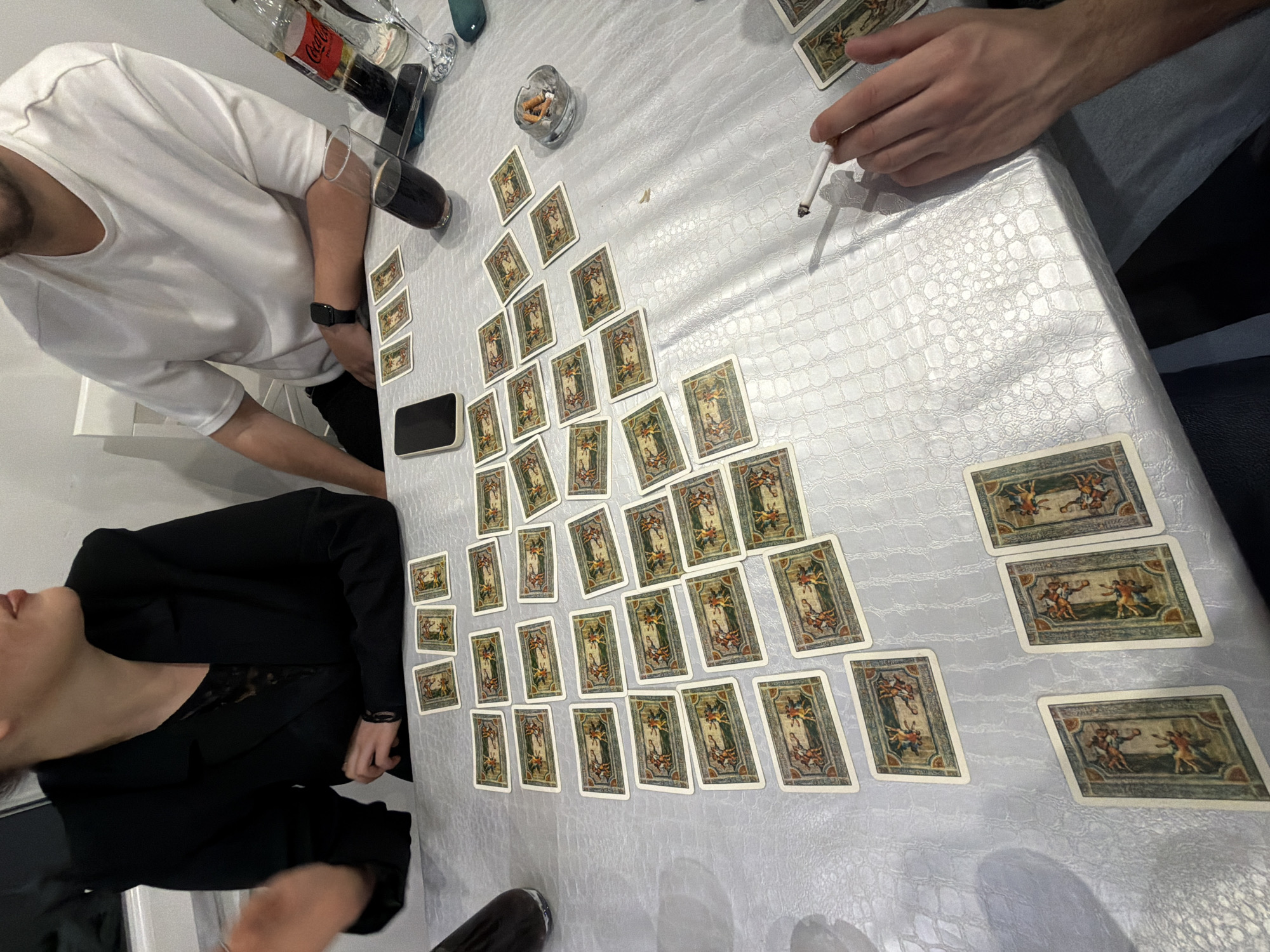 A high-angle shot depicts a group of people around a table covered in a white, textured tablecloth. Spread across the table are numerous playing cards, arranged in a pattern resembling a pyramid or a branching tree. To the left of the frame, a person in a black blazer is partially visible. Another person, wearing a white t-shirt, is positioned near the top left corner, next to a smartphone and a glass of dark liquid, possibly Coca-Cola. In the upper center, there's a small glass ashtray filled with cigarette butts. A hand holding a lit cigarette is visible on the upper right. A second person is seen on the upper right with denim attire. The location cannot be determined.