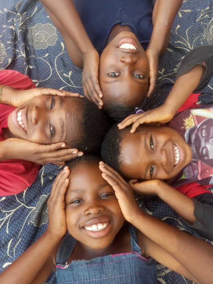 The image presents four children, likely young, arranged in a circle with their heads touching in the center. They are lying down on a dark blue or black fabric adorned with a repeating light-colored, possibly floral or abstract, pattern. All four children are looking directly up at the camera with wide, joyful smiles, revealing their teeth. Their hands are playfully placed around their own heads or on the heads of the children next to them.
The child at the top wears a dark blue top. The child on the left is in a red t-shirt. The child on the right wears a red shirt featuring a graphic design, possibly a helmet. The child at the bottom is dressed in a blue denim-like top or dress with frayed straps. The lighting is bright and even, suggesting the photograph was taken during daytime. The overall impression is one of happiness, playfulness, and camaraderie among the children. There is no visible text in the image. The scene is likely set in a domestic or relaxed outdoor environment in Tempure, Nigeria.