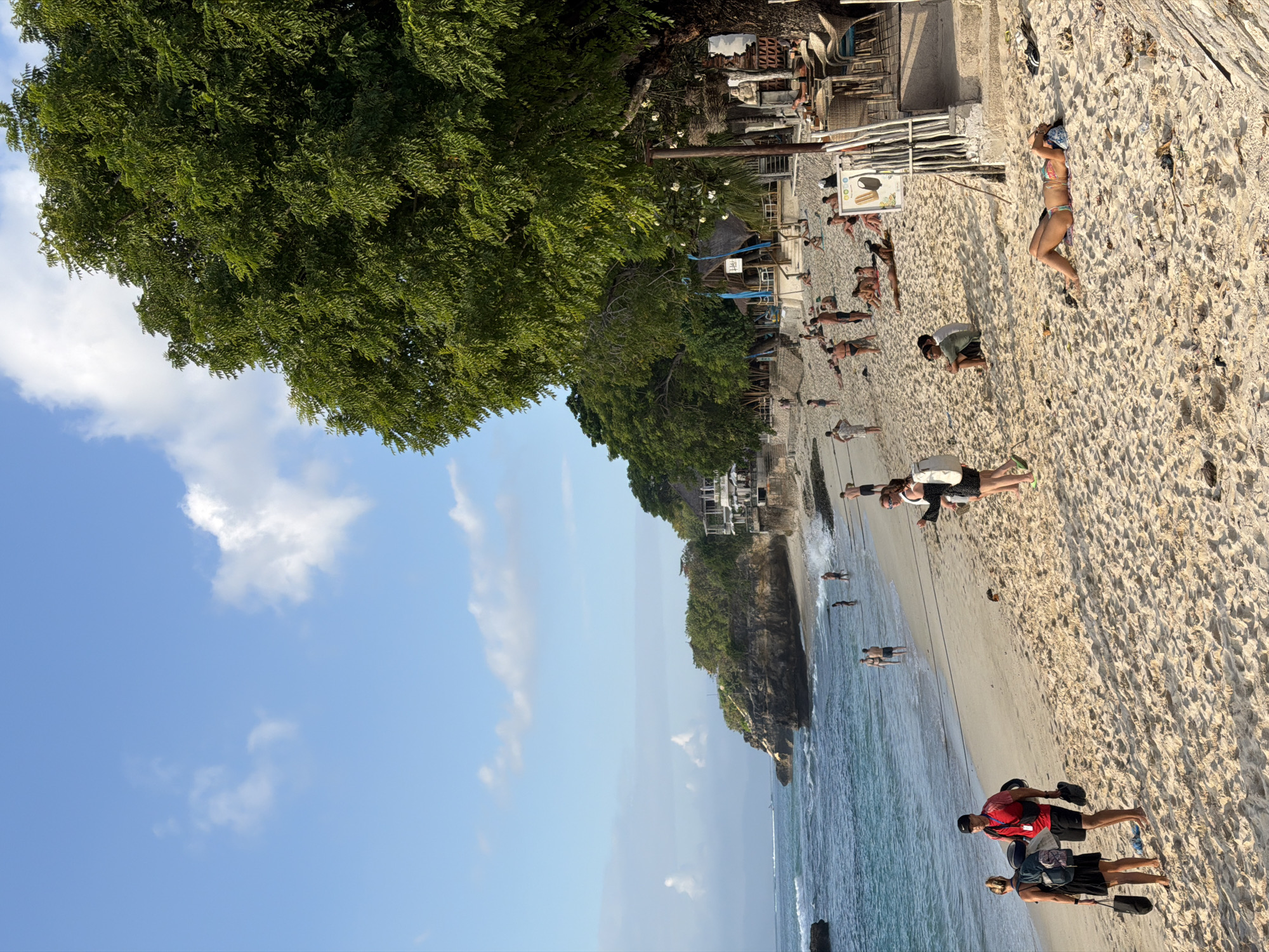 The image depicts a crowded beach scene on Nusa Penida, Indonesia. The sandy beach is populated with numerous people enjoying the sun and sea. In the foreground, two individuals are walking on the sand, while further along the beach, others are swimming or wading in the ocean. Several people are seen sunbathing on the sand.
To the right of the image, a large tree with dense green foliage dominates the scene, providing shade over a beachfront area with tables and chairs, suggesting a cafe or restaurant. Beyond the beach, a cliffside with a building perched on top is visible, with lush vegetation covering the area. The ocean is a clear blue, with gentle waves lapping against the shore. The sky is bright blue with scattered white clouds, indicating a clear, sunny day. The overall impression is one of a vibrant and popular tourist destination.